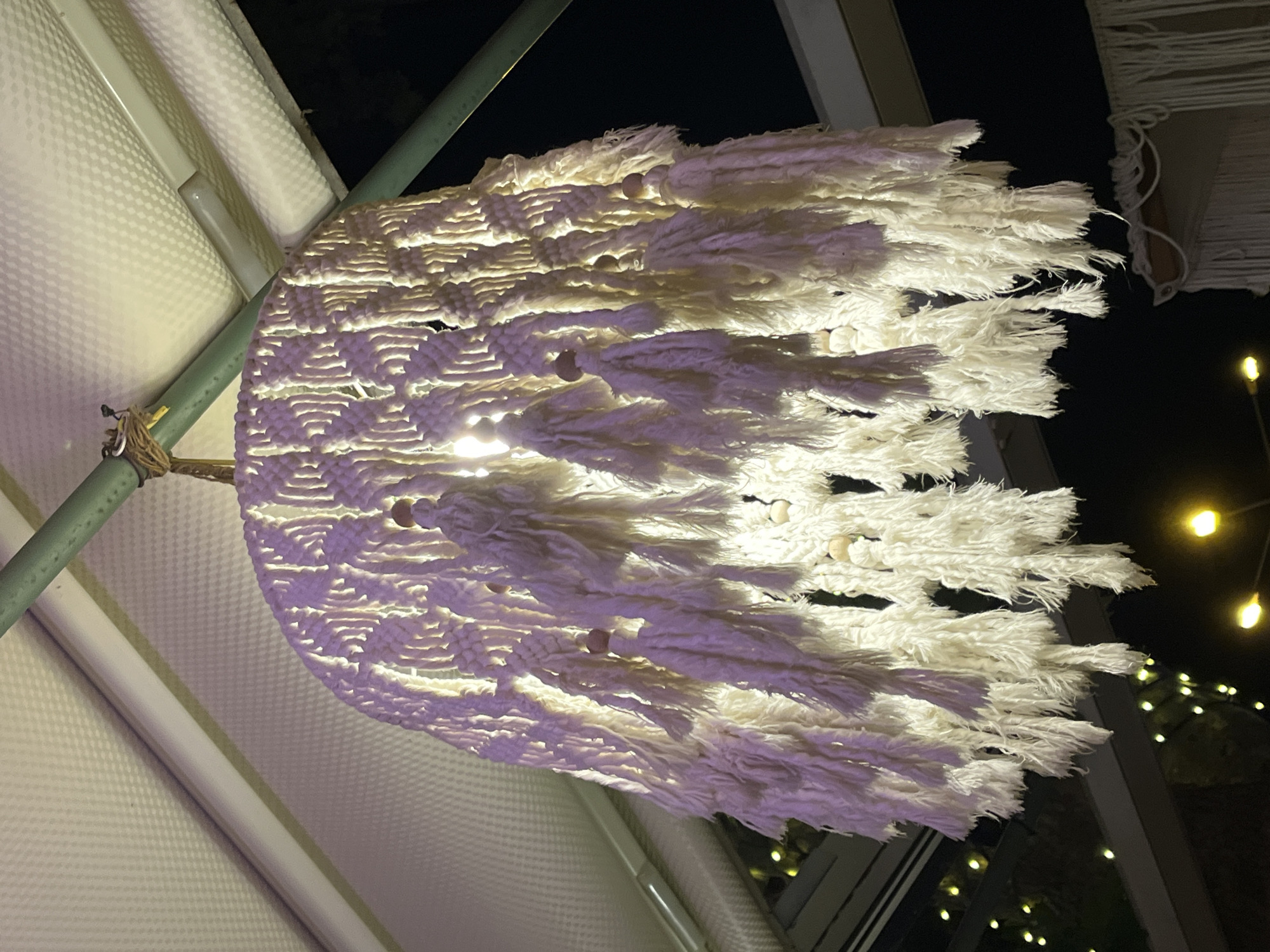 The media file captures an illuminated macrame-style hanging decoration or light fixture at night. The central subject is a rounded object crafted from white macrame cords, featuring intricate knots and numerous fluffy, textured tassels extending outwards. Small, light-colored wooden beads are interspersed throughout the macrame design. A light source within the object casts a bright, warm glow, highlighting the textures and patterns of the woven material and creating a luminous effect.
The object is suspended by a light brown rope wrapped around a green metal pole, suggesting it is part of an outdoor structure, possibly a pergola, gazebo, or canopy. In the background, sections of a white, possibly translucent or patterned fabric are visible, supported by further metal framework. The overall background is dark, indicating nighttime. Out of focus in the distance, numerous small, yellow-orange lights, likely decorative string lights, contribute to a warm and inviting ambiance.
There are no people visible, nor any specific activity taking place beyond the presentation of this decorative lighting. The time of day is clearly night due to the darkness and artificial illumination. The setting, with its outdoor structure and decorative lights, implies a festive, relaxed, or elegant evening atmosphere, possibly in a resort, restaurant, or event space, consistent with the Alanya, Turkey context. No visible text is present in the image.