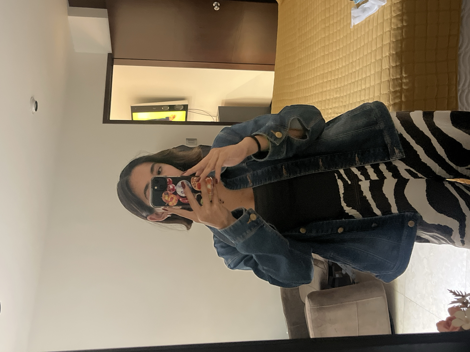 The image shows a woman taking a selfie in a mirror. She is wearing a denim jacket over a black top and zebra-striped pants. The background reveals a hotel room interior, featuring a bed with a quilted gold cover, a mounted television, and a closet. Based on the prompt, the location is presumed to be San Bernardino Tlaxcalancingo, Mexico.