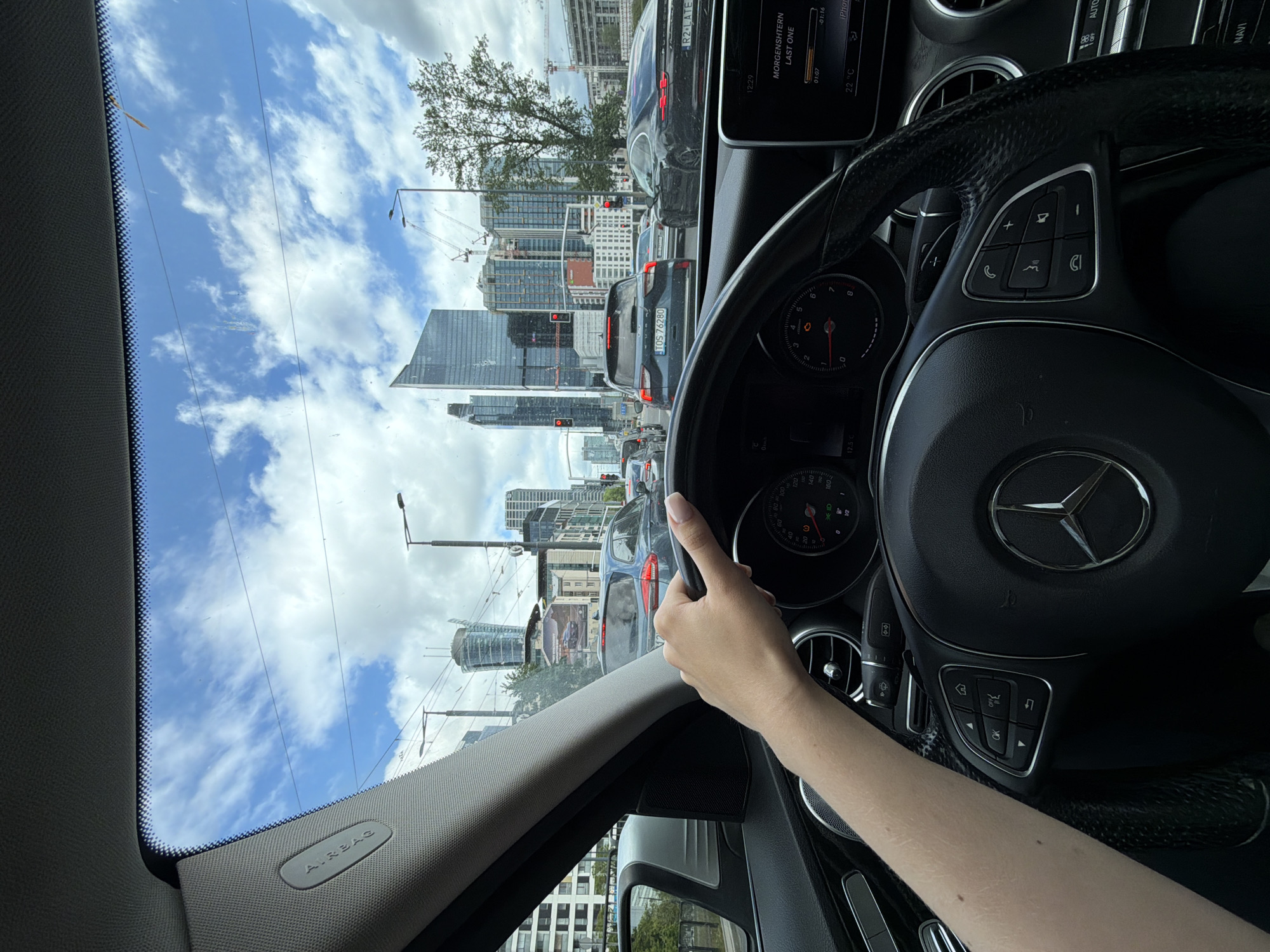 The view from inside a Mercedes-Benz C-Class sedan, showing a driver's hand on the steering wheel. The dashboard is visible, with the instrument cluster, infotainment screen, and controls. The car is in heavy traffic. Skyscrapers are visible in the distance through the windshield. The car is in Warsaw, Poland, as evidenced by the registration plates of the vehicles.