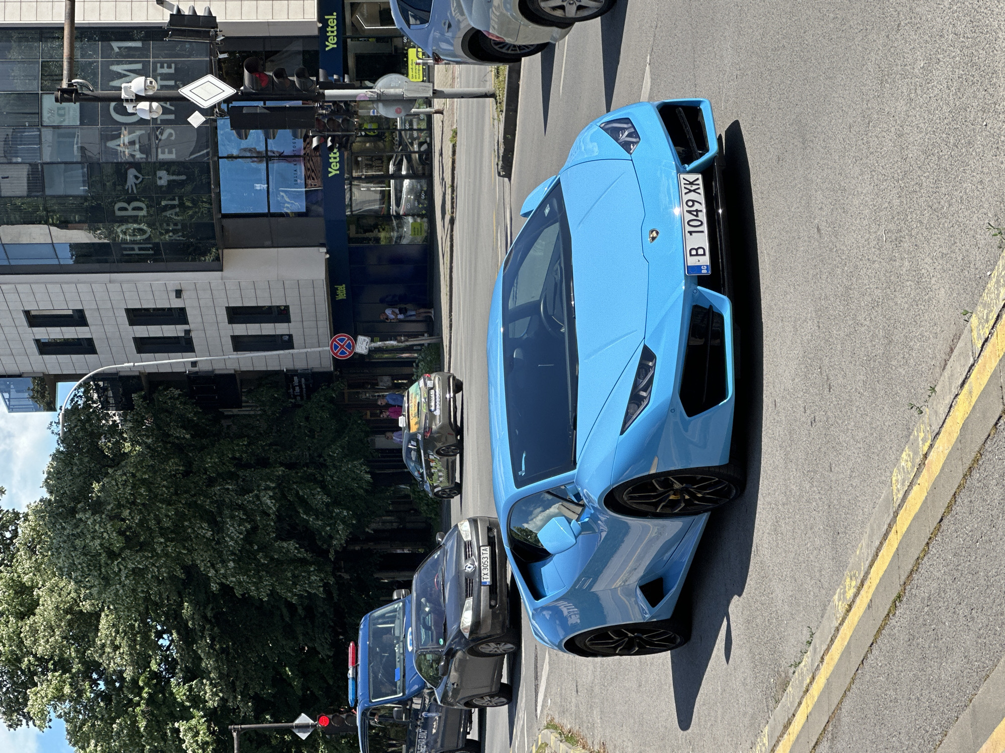 The image shows a light blue Lamborghini parked on a street with a double yellow line marking near the curb. In the background, there are several other vehicles and a building with a glass facade displaying the name "HOB GM1 REAL ESTATE". A street sign can be seen next to the building. In addition, the building has signage for "Vettel" and "Yettel". The license plate on the car is "B 1049 XK". The building is located in Varna, Bulgaria.