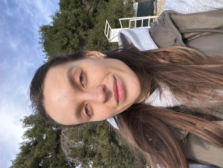 A young adult female is depicted in a selfie-style portrait taken outdoors in Boreti, Montenegro. She has long brown hair, light skin, grey-blue eyes, and a subtle smile, looking directly into the camera. She wears a light grey hooded sweatshirt and an olive green quilted vest. A silver hoop earring is visible in her right ear. The background consists of dense green trees, likely conifers, under a partly cloudy blue sky. To her right, a white, two-tiered structure consistent with a lifeguard stand is partially visible, situated above a light-colored, possibly pebbled ground surface, indicating a coastal or beach environment.