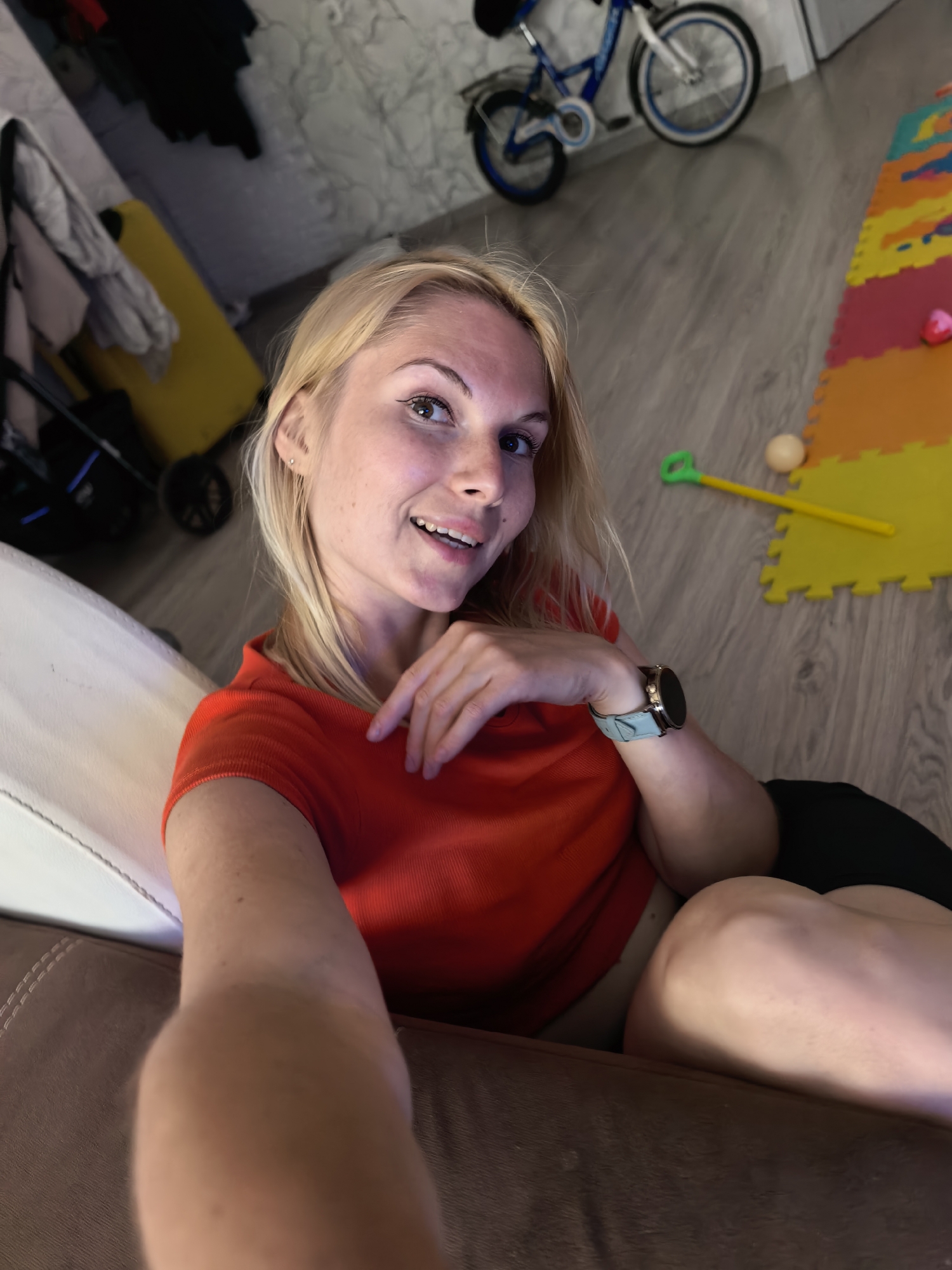 A blonde woman in her 20s is sitting on a brown sofa, taking a selfie. She is wearing a red t-shirt. Her left arm is resting on her lap, and her right hand is on her chest. She is smiling at the camera. In the background, there is a white wall, a yellow suitcase, a blue bicycle, and a colorful play mat with toys. The scene appears to be an indoor setting, possibly a living room or playroom. The lighting suggests it is daytime or the room is well-lit. There are no discernible landmarks or elements that would specifically place this image in Sterlitamak, Russia.