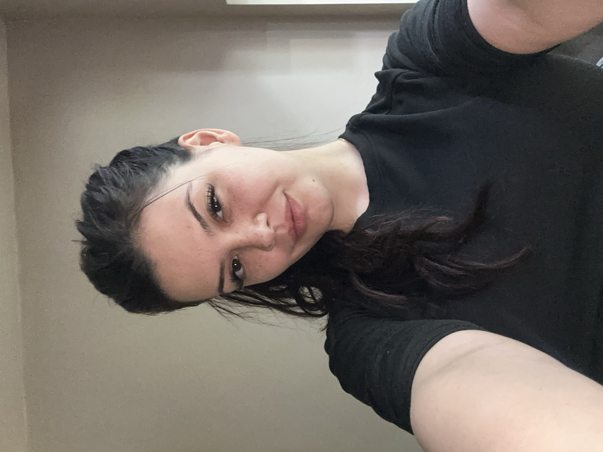 A head-and-shoulders selfie photograph shows a young woman with dark hair and light skin. She is wearing a dark t-shirt. The background consists of a light beige wall and a small section of white trim near the top. The photograph's orientation is such that the woman's face is tilted towards the right. The location is not discernible from the provided image.