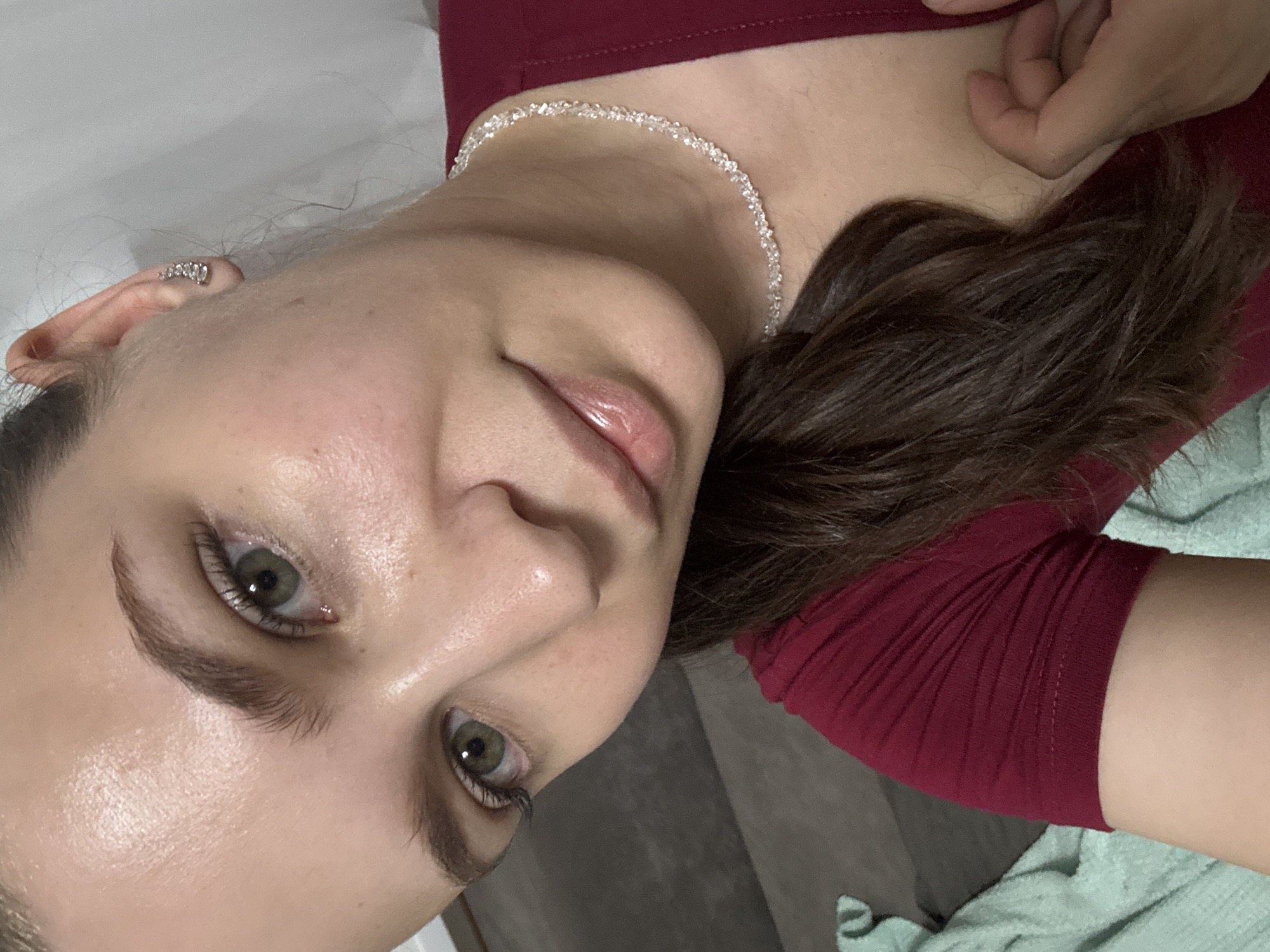 The image shows a close-up, rotated view of a person's face and upper body. The subject has light skin, green eyes, and dark brown hair. They are wearing a maroon-colored shirt and a delicate, sparkly necklace. A silver earring is visible on their left ear. The lighting suggests an indoor setting, possibly natural light, given the subtle highlights on the skin. The background is blurred, showing hints of a gray and white environment, with a light blue fabric visible towards the bottom right. The person's expression is neutral to slightly curious. There is no visible text in the image. The overall impression is a personal, casual photograph.