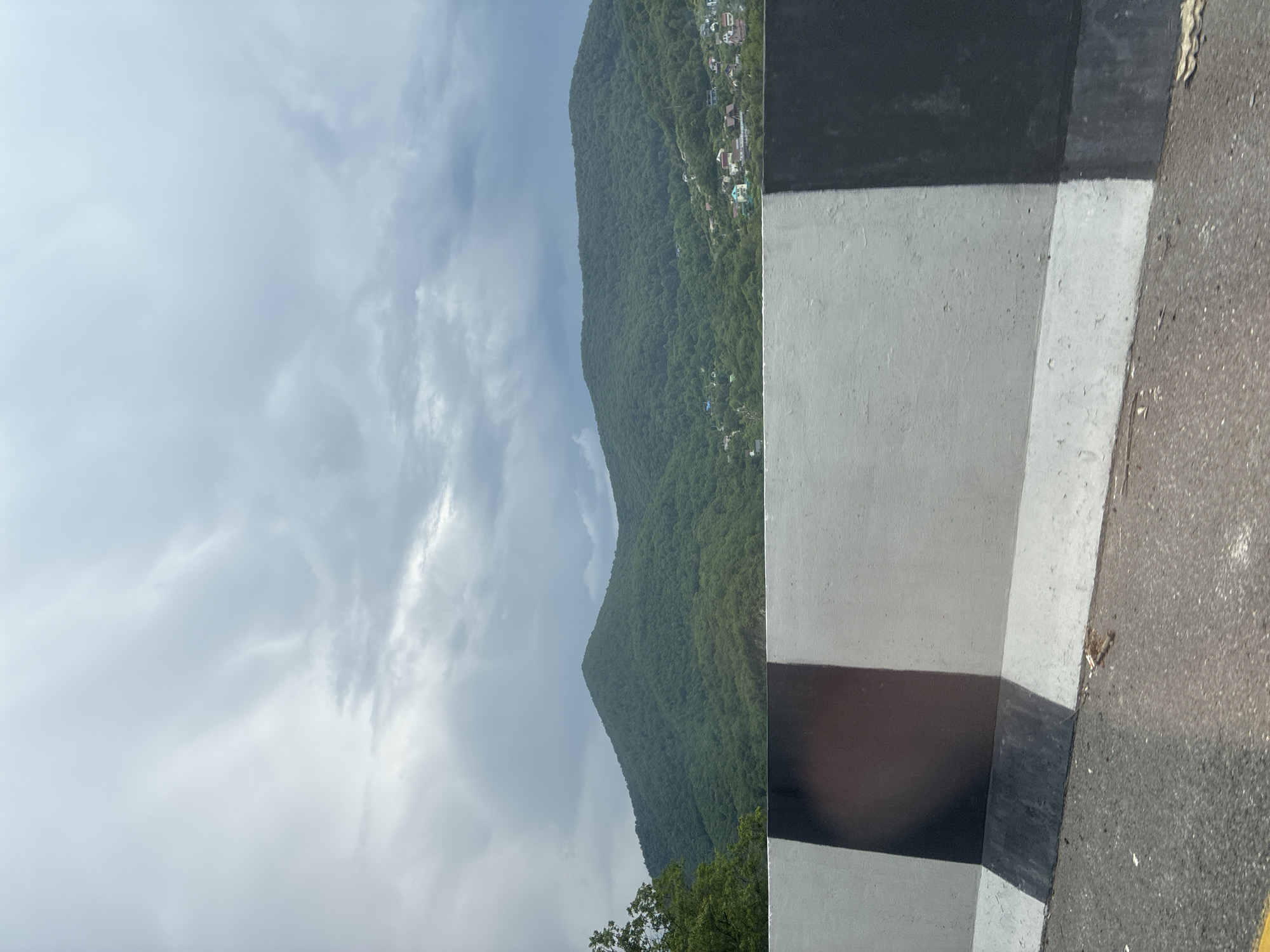 The image depicts a roadside view with a concrete barrier in the foreground and lush green mountains in the background. The sky is overcast with heavy clouds. The barrier is painted with alternating black and white stripes. Scattered houses and buildings are visible nestled on the slopes of the mountains. The scene appears to be a natural landscape with some human habitation, possibly a rural or suburban area. The location is identified as Dederkoi, Russia. The image composition suggests the photo was taken from a moving vehicle, as indicated by the angle and the slight blur on the roadside. There are no people or animals visible in the frame.