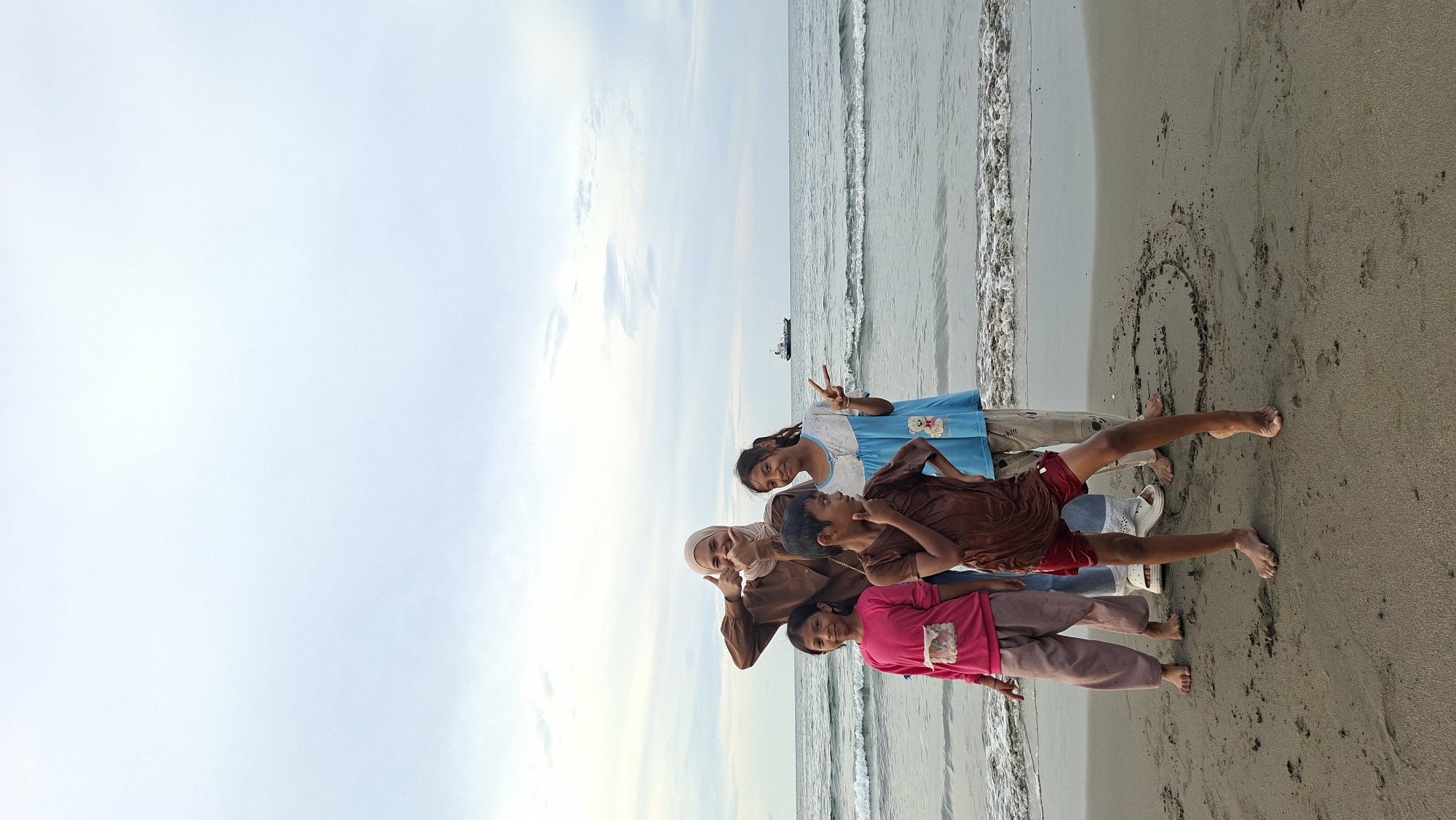 An outdoor, full-shot, eye-level photograph captures a group of four individuals on a beach in Lhokseumawe, Indonesia, with the sea and sky in the background. The individuals, appearing to be two adults and two children, are positioned on the wet sand near the shoreline.
In the front, a child, possibly male, wearing a wet, dark brown t-shirt and red shorts, stands with his left leg bent and foot elevated. His right hand is near his chin, and he is looking to the right. To his left, another child, possibly female, in a bright pink long-sleeved shirt and light brown pants, stands with her arms down by her sides and looks towards the camera. Behind these two children, another child, possibly female, in a light blue t-shirt with cartoon graphics and beige pants, is striking a peace sign with her right hand and looking to her left. Behind her, a person wearing a brown hijab and brown long-sleeved shirt is giving a thumbs-up gesture with both hands and smiling, looking at the camera. The group is gathered on a sandy beach with gentle waves lapping at the shore. In the distance, a ship is visible on the horizon. The sky is overcast with a light gray hue.