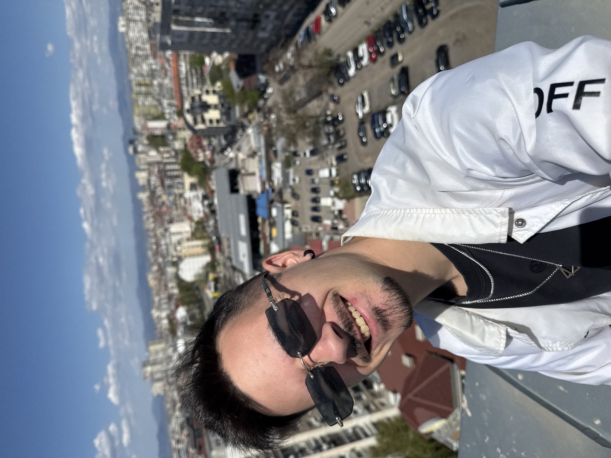 A man with sunglasses stands on a rooftop overlooking Sofia, Bulgaria. He has dark hair, a light mustache, and is wearing a white jacket over a black shirt with a silver necklace. The jacket reads "OFF". The city is visible in the background, with buildings, a parking lot full of cars, and a blue sky with clouds. The photograph is taken at an angle, with the man's face in the foreground and the city in the background.
