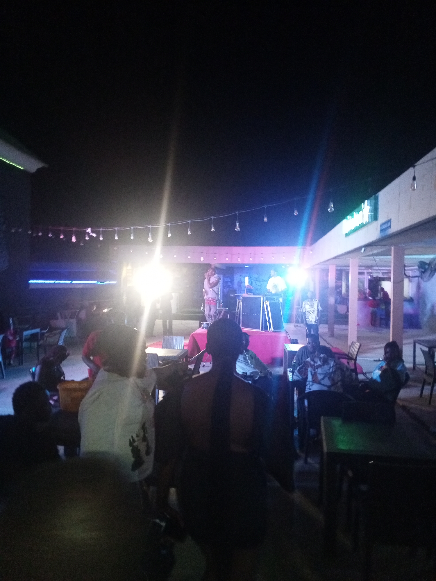 The scene captures an outdoor nightlife setting in an unidentified location. A brightly lit stage features a person with a microphone, possibly performing. A DJ station is set up near the performer. There are tables and chairs spread around the area, occupied by patrons. The backdrop includes strung lights and signage on the building facade. A mix of clothing is worn by people, including t-shirts and button-down shirts. Interactions are suggested through seating arrangements around tables, though the low light and distance make specific interactions difficult to define.