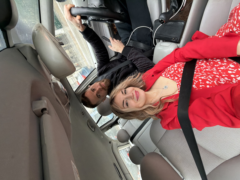 The image depicts two individuals inside the cabin of a light-colored vehicle, presumed to be in Budva, Montenegro. A female is positioned in the front passenger seat, while a male occupies the driver's seat.
The female, with long wavy blonde hair, is wearing a red blazer over a red dress with a white floral pattern. She has a silver necklace with a circular pendant and is smiling directly at the camera. Her seatbelt is fastened across her chest.
The male, with short brown hair and a beard, is wearing a black quilted jacket. His right hand is on the steering wheel, and his left hand holds a black smartphone. He is looking towards the camera with a faint smile. His seatbelt is also fastened.
The car's interior features light grey fabric seats, a matching headliner, and sun visors. A black bag or object rests on the center console between the seats, near the gear shift lever. Through the front and side windows, a blurred urban environment is visible, featuring buildings and other vehicles, with some indications of wet conditions on the glass. The rearview mirror is also visible.