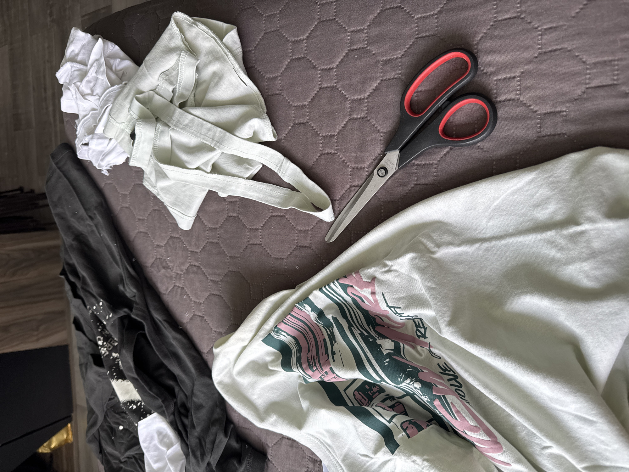The image presents an overhead view of various clothing items and a pair of scissors laid out on a dark brown or grey quilted surface, possibly a bed or mattress protector featuring a hexagonal pattern.
In the center, a pair of black-handled scissors with distinctive red finger loops lies open, pointing towards the top right. The silver blade of the scissors is marked with the words "SOLINGEN" and "GERMANY." To the right, a white t-shirt is spread out, showcasing a prominent, abstract graphic design in shades of pink and dark green/teal. Partially visible text, including letters like "RED" or "RE," is integrated into this design.
On the left side of the frame, near the scissors, a light green or pale yellow fabric item with straps, resembling a tank top or cut-up cloth, is visible. Adjacent to this, a crumpled pile of white fabric, likely textile scraps or a garment, is gathered. Further to the far left, a stack of dark-colored garments, predominantly black or dark grey t-shirts with some white splatter patterns, is partially obscured.
The overall scene suggests an activity involving fabric and cutting, such as a craft project, repurposing old clothes, or preparing materials. The setting is indoors, likely a domestic space, with a wooden floor and a dark piece of furniture visible in the background on the far left. The lighting appears natural, indicating daytime. No individuals are present, and no specific emotions are conveyed. The location context provided for this image is San Bernardino Tlaxcalancingo, Mexico.