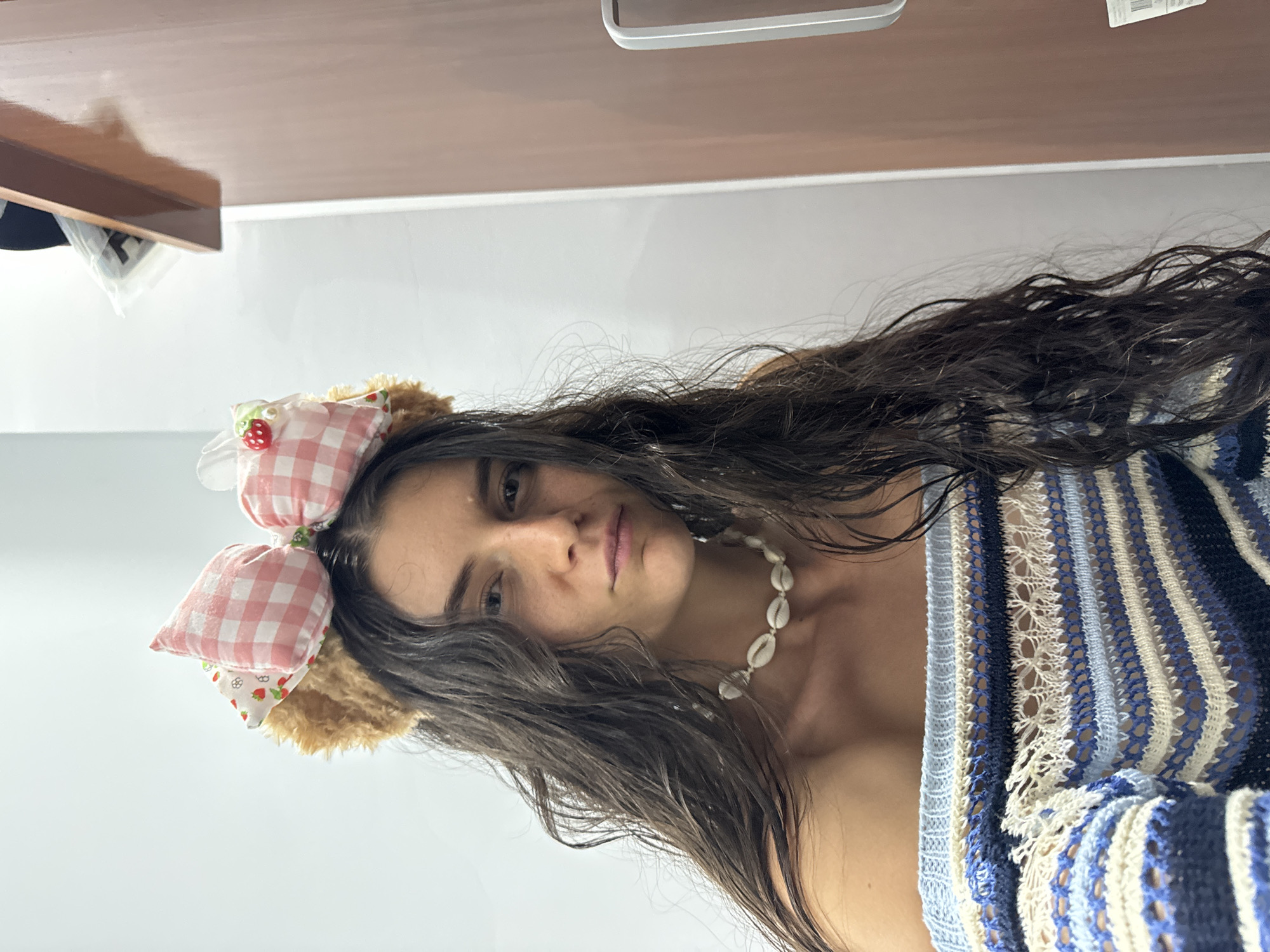 A close-up portrait of a young woman is presented, rotated 90 degrees clockwise. The woman has long, dark, wavy hair that appears damp. She wears a white seashell necklace around her neck and a blue, white, and dark blue striped, open-knit or crocheted top. On her head, she has a whimsical headband featuring two puffy pink-and-white gingham bows, one of which has a small red strawberry embellishment. The headband also includes furry brown sections, giving the appearance of animal ears.
Her face is turned towards the camera, and she has a neutral to slightly contemplative expression with slightly parted lips. The background shows a plain white wall topped by a reflective, light brown or wooden surface, possibly a shelf or desk. A crumpled plastic bag is visible beneath the brown surface on the left side of the frame, and a silver-colored object can be seen in the upper background. The setting appears to be indoors. No specific activity beyond posing for the camera is depicted, and no text is visible in the image. The location context provided is Mũi Né, Vietnam.