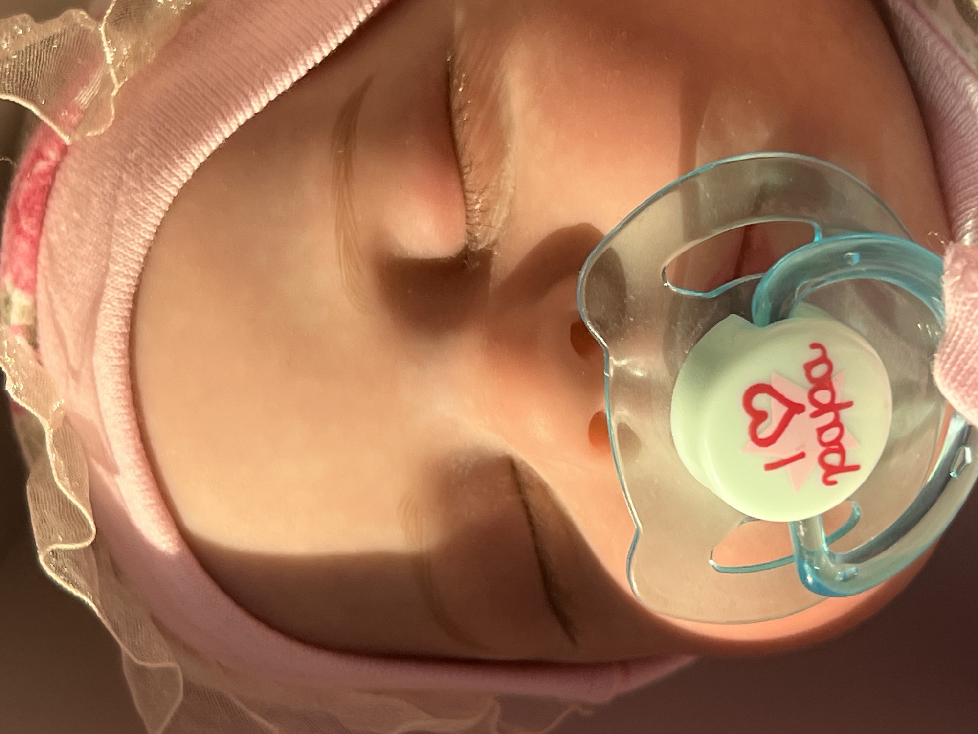 This is a close-up, eye-level, vertical photograph of a sleeping baby wearing a pink headband with lace trim. The baby has closed eyes and a pacifier in its mouth. The pacifier is clear plastic with a light green nipple shield that has a pink heart and the words "I love papa" written in red. The baby's face is illuminated by warm, directional light that casts a shadow across its forehead. The background is out of focus and appears to be a soft, neutral color. The image does not provide any information about the location in Votkinsk, Russia.