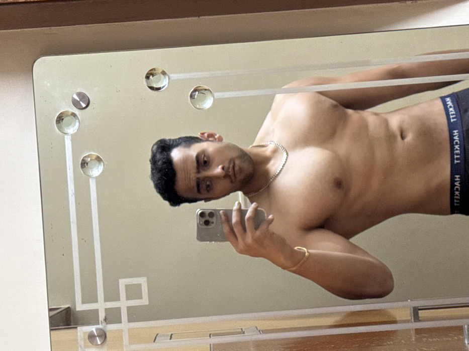 A young man, likely of Indian descent, is captured in a mirror selfie within an indoor setting, possibly a bedroom or bathroom, in Kolkata, India. He is shirtless, showcasing a muscular physique with defined chest and abdominal muscles, and wears dark-colored underwear. The elastic waistband of his underwear displays the mirrored text "HVCKELL" (likely "HACKELL").
He holds a modern smartphone in his left hand to take the picture, his gaze directed confidently towards the camera's reflection. Around his neck, he wears a silver-colored chain, and a gold-colored bracelet adorns his left wrist. The mirror itself is decorative, featuring frosted glass patterns and several circular, crystal-like embellishments along its edges, some secured by metallic studs. The background reflected in the mirror appears to be a plain, light-colored wall. The overall image presents a posed shot emphasizing the man's physical form.