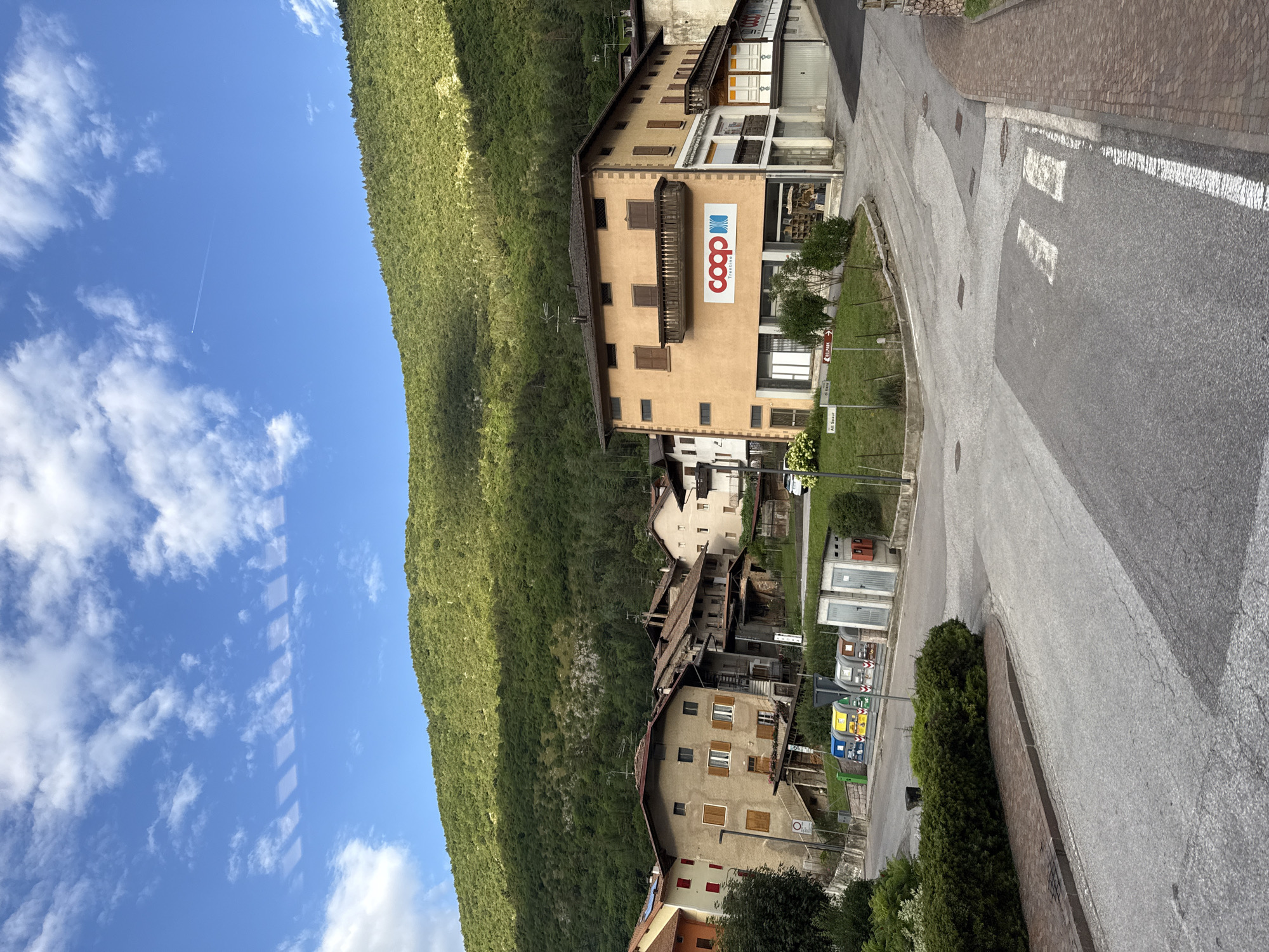 The image depicts a street scene in Spormaggiore, Italy. The perspective is from a slightly elevated position, looking down a winding road. On the left side of the road, a steep, heavily forested mountainside dominates the scene, with a bright blue sky dotted with white clouds above it. A faint contrail from an airplane is visible in the sky.
The right side of the road features a cluster of buildings. The most prominent building is a multi-story structure with a light orange facade, featuring several windows and balconies. A large, red and white "coop Trentino" logo is prominently displayed on this building, indicating a retail cooperative. Below this building, other structures are visible, suggesting a small town or village. The road itself is paved, with white pedestrian crossing markings and various signs. To the far right, a brick-paved sidewalk runs alongside the road.
In the foreground, there is a green grassy area with some bushes and trees. There are also several bins for waste separation visible near the buildings. The overall impression is of a quiet, picturesque mountain village on a clear day. The time of day appears to be daytime, likely late morning or early afternoon, given the bright sunlight and shadows. There are no people visible in the image.