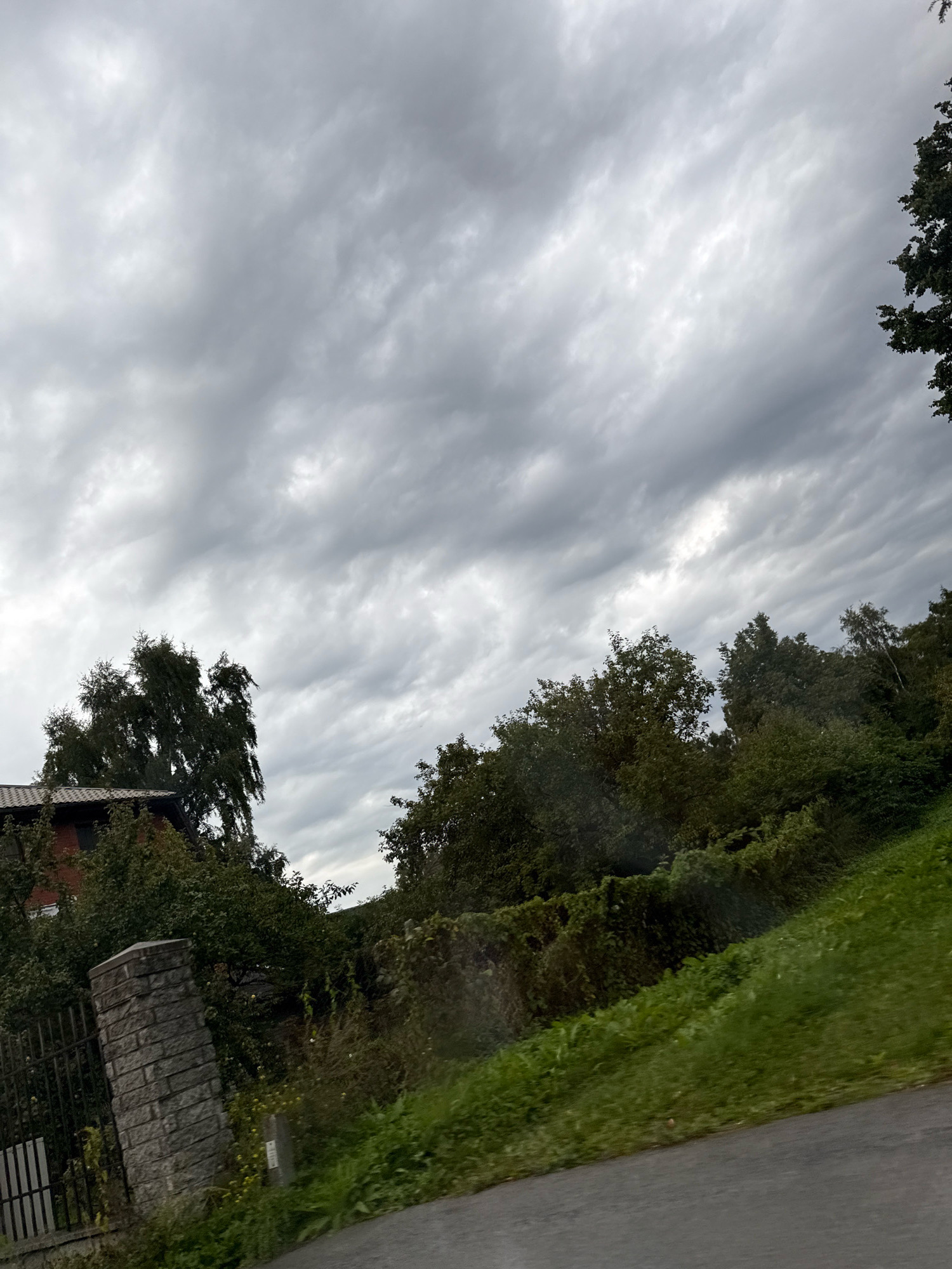 The image depicts a cloudy sky over a residential area in Tallinn, Estonia. In the foreground, a paved road curves to the right. To the left of the road, a low stone wall marks the edge of a property, with a dark metal fence behind it. A red-bricked building, partially obscured by trees, is visible behind the wall. Lush green vegetation, including trees and bushes, dominates the middle ground. The sky is filled with a dense layer of grey clouds, suggesting overcast weather. No people are present in the image.