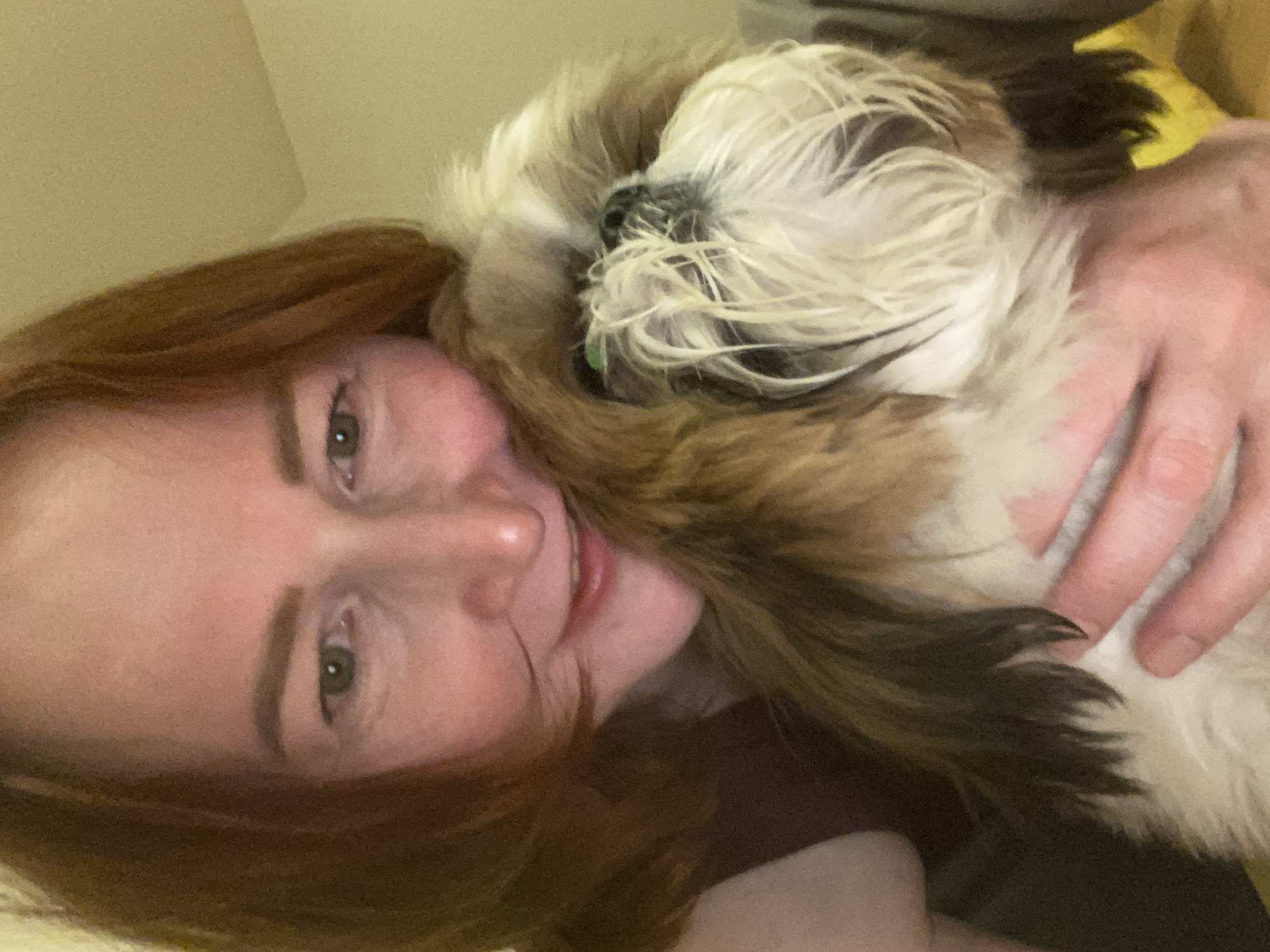 A selfie photograph depicts a person with auburn hair, green eyes, and fair skin, smiling and holding a Shih Tzu dog. The dog has a mix of white and brown fur. A hand is visible holding the dog. The setting appears to be indoors, likely a residential space, with a neutral-colored wall in the background. No evidence indicates the location is in Chaikovsky, Russia.