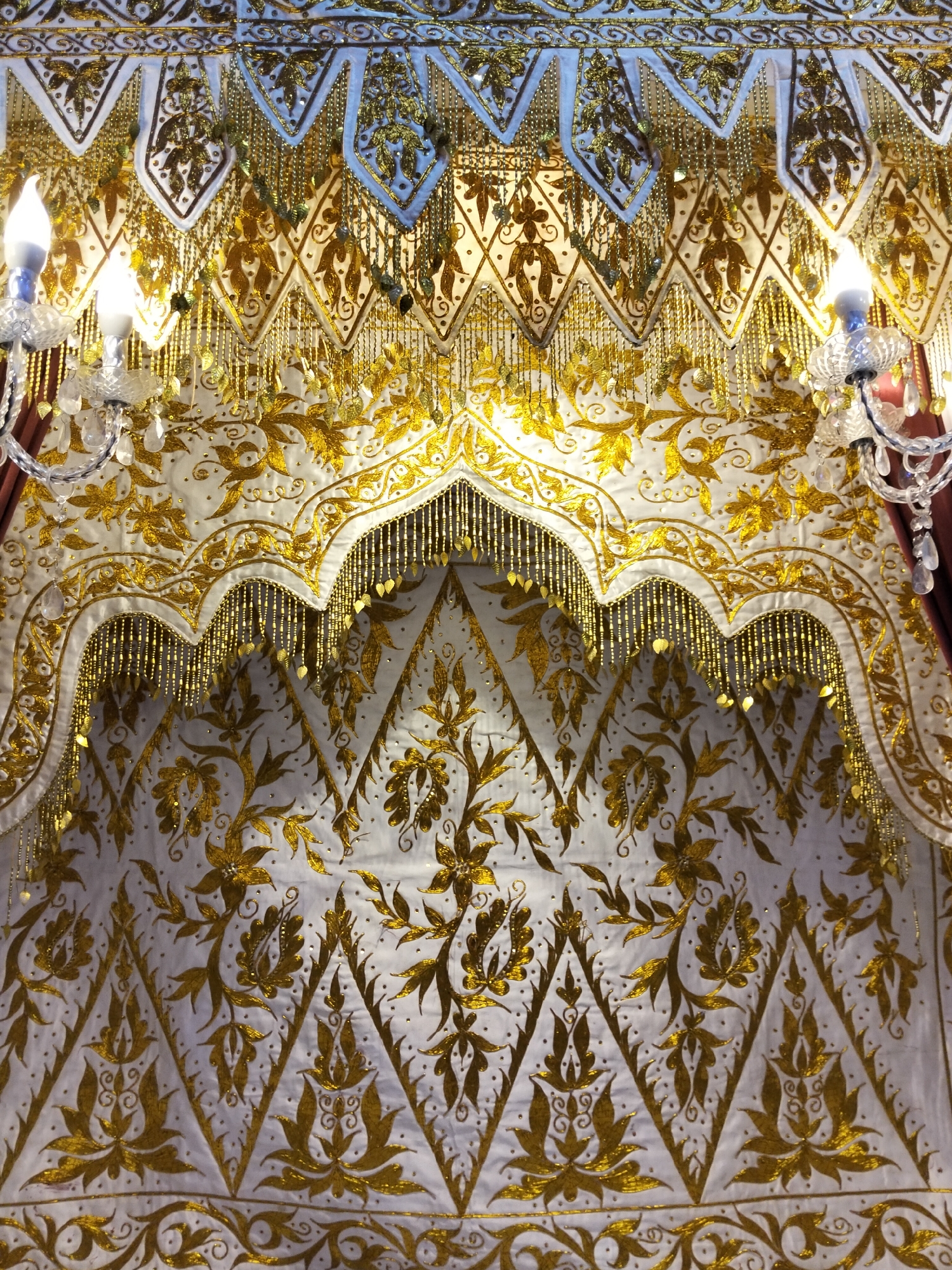 The image shows a close-up of an ornate, white fabric with extensive gold embroidery. The embroidery features floral and leaf motifs arranged in a repeating pattern. Decorative gold bead fringe hangs from scalloped edges in the upper portion of the fabric. Two crystal sconces with lit bulbs are visible on either side of the frame, attached to what appears to be a curtain rod or decorative fixture. The overall impression is one of richness and intricate craftsmanship. The location, Cottrueng, Indonesia, is noted as context but is not visually represented in the image itself.