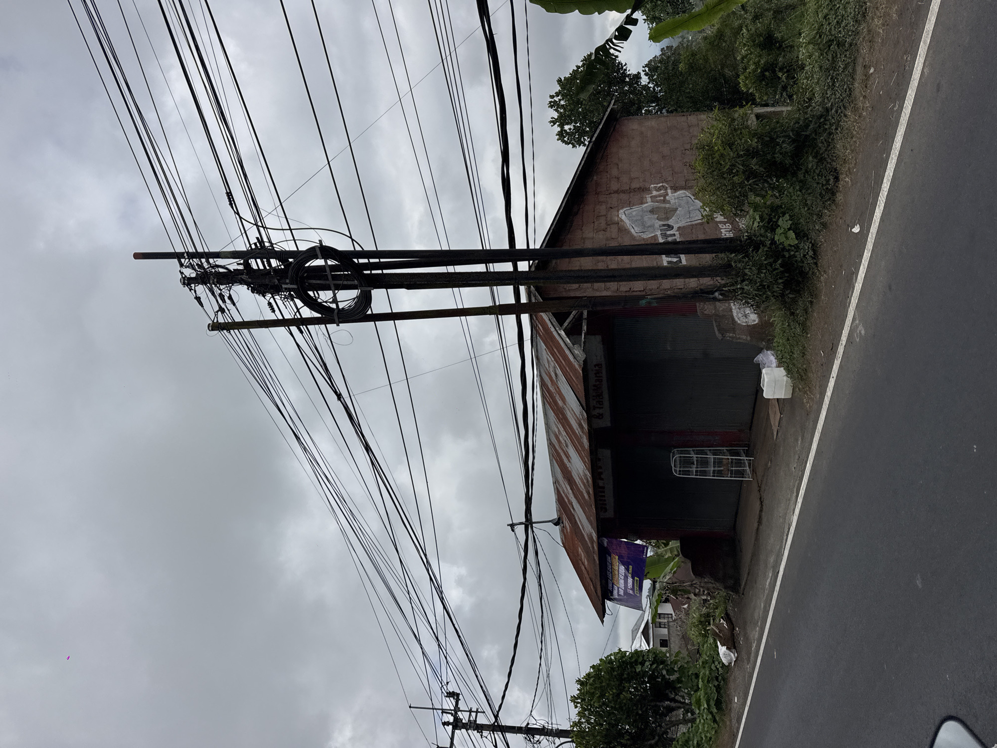 The image depicts a roadside scene in Kintamani, Indonesia, characterized by a cloudy sky and an abundance of electrical wires overhead. In the foreground, a gray asphalt road runs along the bottom of the frame, with a white dashed line marking its edge. Adjacent to the road is a slightly elevated dirt embankment.
Dominating the midground is a small, weathered building with a corrugated metal roof that appears to be rusted. The building has large, closed metal shutters, suggesting it is a shop or commercial establishment. The front of the building is constructed from red brick and has some graffiti or painted markings on its wall, including the text "KARTU AS" in large white letters, with a heart shape above the "U". A tall, dark electrical pole with a complex network of wires and insulators stands prominently next to the building, contributing to the cluttered overhead electrical infrastructure. To the left of the building, a white metal shelving unit is visible near the entrance.
Behind the building, lush green vegetation and trees are present. In the background, several houses with white walls and tiled roofs are visible, indicating a residential area. The overall environment suggests a rural or semi-urban setting in Kintamani, likely along a main road given the presence of the electrical infrastructure and the road itself. No people are visible in the image. The atmosphere is somber due to the overcast sky.