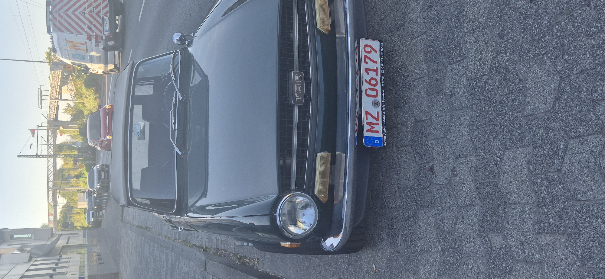 A dark green Triumph TR6 convertible is parked on a cobblestone street in Mainz, Germany. The car is facing forward, with its front grille and headlights visible. The license plate reads "MZ 06179" and includes "D" for Deutschland, "Avino Automobile & Kfz-Service Mainz," and a phone number. The street is lined with a building on the left and other vehicles in the background, including a red car and a white van with an orange chevron pattern. An elevated railway bridge can be seen in the distance. The scene appears to be in an urban area, likely a street in the city of Mainz.