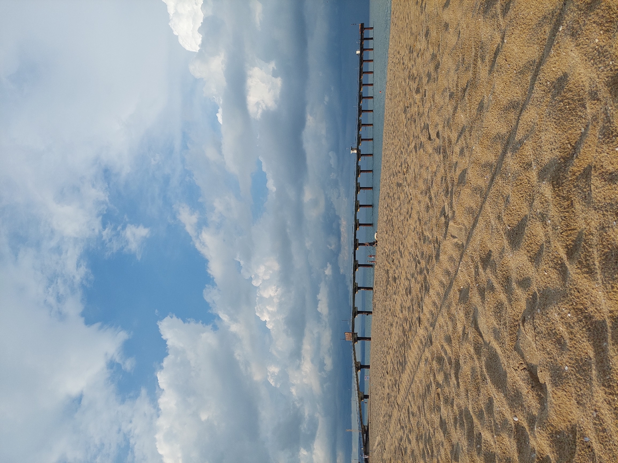 The image shows a view of the beach and the sea at Shkorpilovtsi, Bulgaria. The foreground is dominated by sand dunes with footprints. In the middle ground, a long pier extends out over the sea. The pier is supported by numerous pillars and has a walkway on top. There are a few small figures of people visible on the pier. The sea is calm and a light blue. The sky above is filled with dramatic clouds, a mix of white cumulus clouds and darker, more ominous storm clouds. The overall impression is one of a picturesque, yet slightly brooding, coastal landscape.