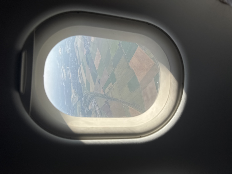An airplane window frames a patchwork of agricultural fields below, seen from above. The fields are predominantly green and brown, suggesting cultivated land. A road or river winds through the landscape. The view is slightly hazy, possibly due to altitude or atmospheric conditions. Sunlight streams onto the window frame, creating bright highlights and deep shadows, adding depth to the composition.