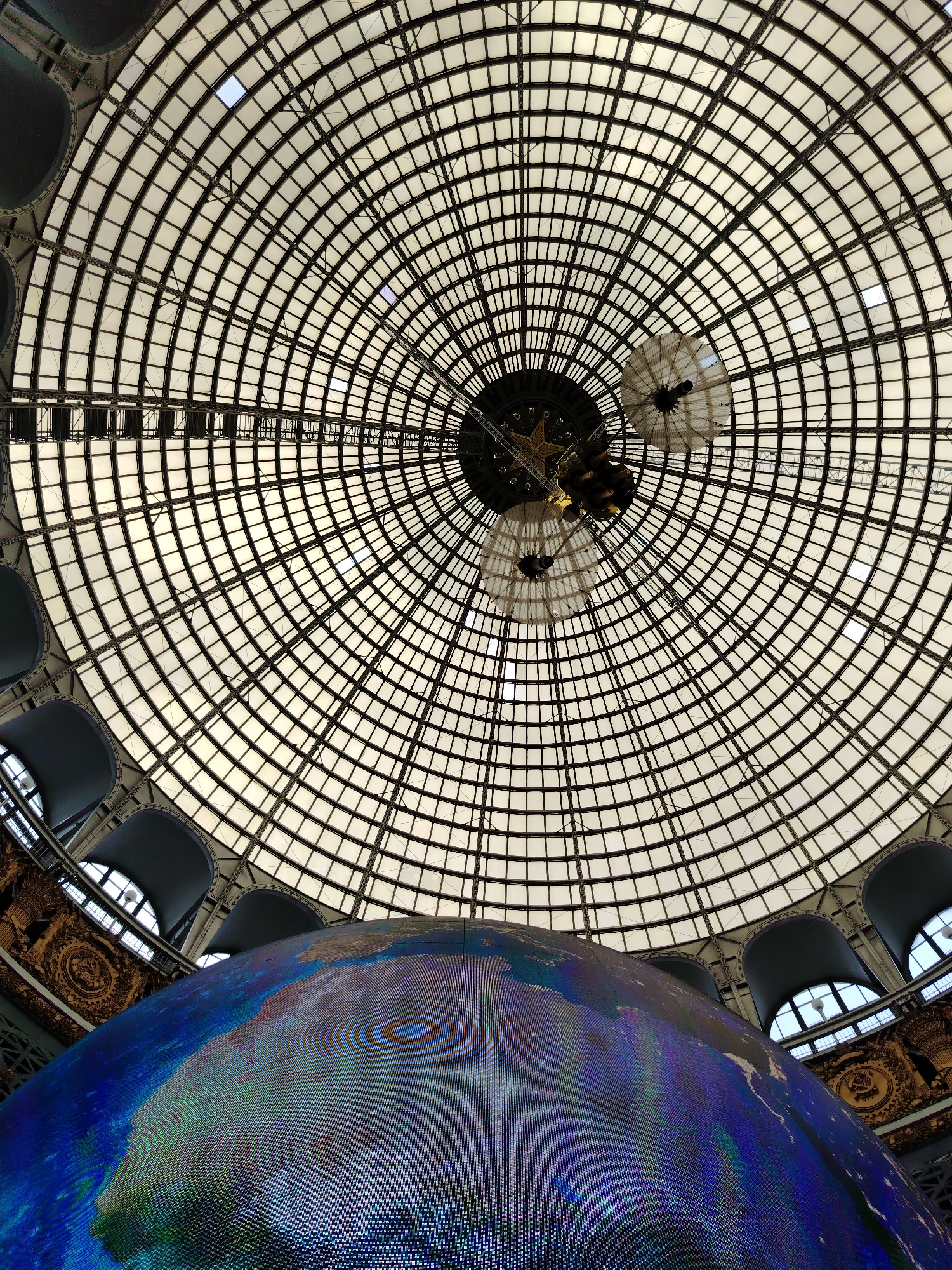 The image depicts the interior of the State Museum of Cosmonautics in Moscow, Russia. The focal point is a large, illuminated model of Earth, rendered with a pixelated effect, occupying the lower portion of the frame. Above the Earth model, a vast, domed ceiling is visible, constructed with a grid of translucent panels supported by a dark, intricate metal framework. Suspended from the center of the dome is a model of a spacecraft, appearing somewhat indistinct due to the lighting and distance. The overall scene conveys a sense of awe and scientific exploration, with the illuminated globe and the expansive, technologically advanced dome creating a dramatic visual impact. Architectural details, such as arched windows and ornate decorations along the sides of the dome's base, are also discernible.