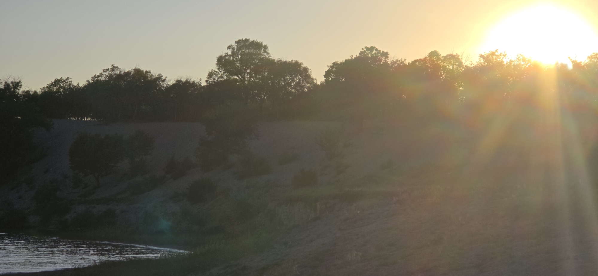 A wide shot shows a bright sun setting over a treeline on a sandy hill. The sun is a large, glowing orb in the upper right corner of the frame, with rays of light fanning out and creating a lens flare effect. Below the treeline, the sand appears to be a light tan color, with some darker patches of vegetation. In the foreground, a body of water, likely a lake or river, reflects the light of the setting sun, creating a shimmering surface. The overall impression is one of a serene natural landscape at dusk. The location is the city of Issyk-Kul, Kyrgyzstan.