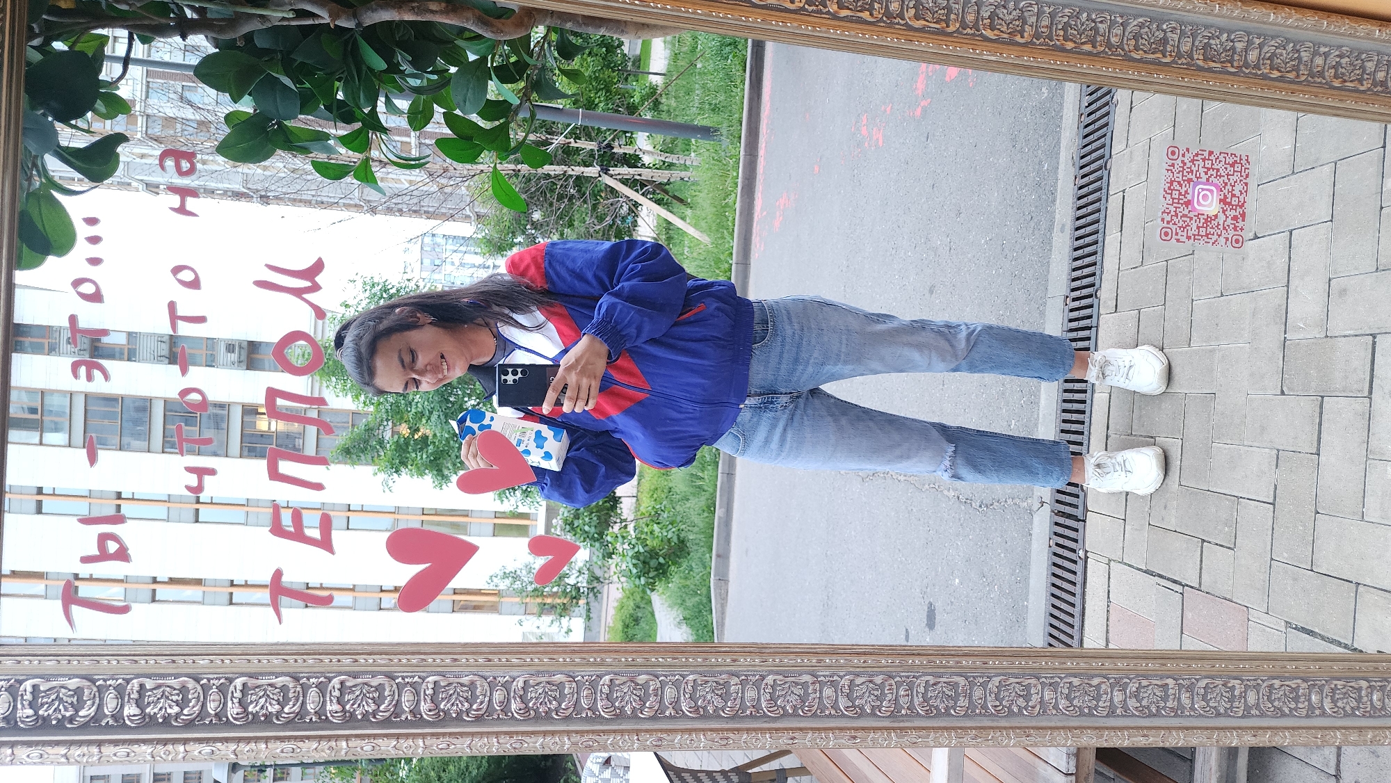 A full-shot, mirrored view of a person outside in what appears to be a city environment. The person, likely female, is standing and holding a carton of milk and a cellphone. The person is wearing jeans, white shoes, and a blue jacket with red detailing. The reflection includes a red heart and red writing (likely Cyrillic).
The background consists of an exterior urban setting: a building facade with numerous windows and general greenery. The mirror is framed by an ornate gold-colored border. On the ground in front of the mirror is a patterned brick surface and a QR code with the Instagram logo. This suggests the location is a public installation designed for taking photos and promoting social media engagement.
It is likely this image was taken in Moscow, Russia based on the Cyrillic writing and the QR code potentially leading to a Russian Instagram page.