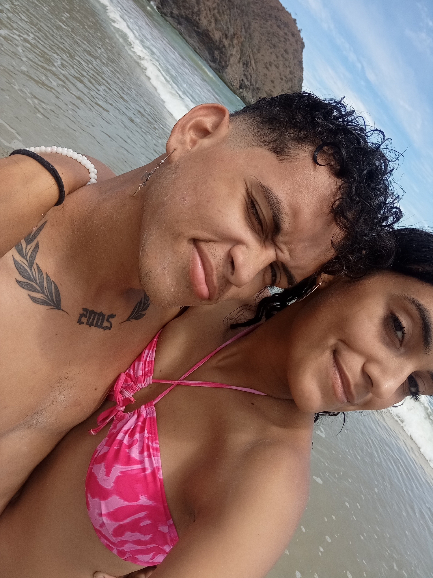A close-up, angled medium shot captures a couple on a beach, likely in or near the water's edge. The man, with curly dark hair, is shirtless and has a visible "2005" tattoo on his chest. He has a black and white beaded bracelet on his left arm and a slender earring on his left ear. His eyes are closed, and he appears to be squinting. The woman, with dark hair, is wearing a pink patterned bikini top and has an earring on her left ear. She is smiling and looking toward the camera. The background includes a calm ocean with small waves, a land formation resembling a rocky hill or small mountain and a cloudy blue sky. The location is Altagracia, Venezuela.