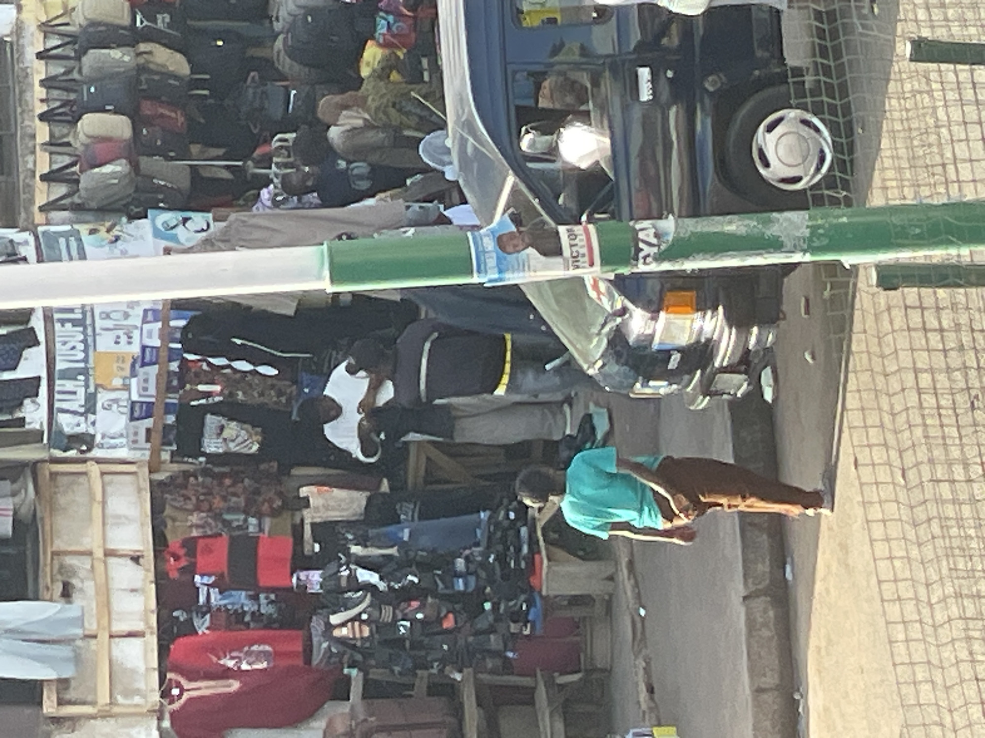 The image captures a bustling street scene in Bukuru, Nigeria. A variety of goods are displayed on makeshift stands, including what appears to be clothing, bags, and possibly shoes. Several people are present, some browsing the merchandise, others standing or walking nearby. A blue vehicle is partially visible on the right side of the frame. The environment suggests an outdoor marketplace with paved ground.