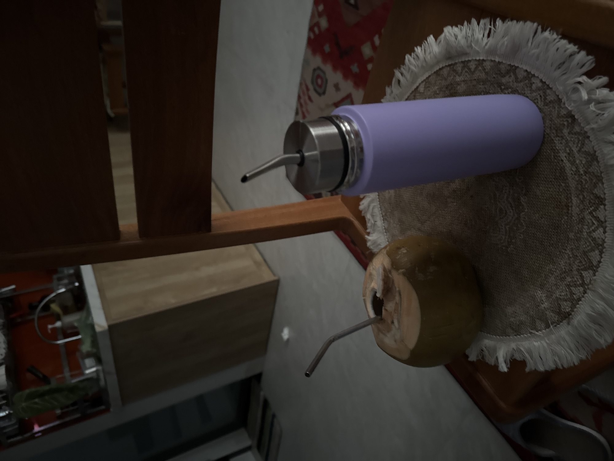 A purple bottle with a silver spout sits on a fringed doily on a wooden chair. A coconut with a metal straw also rests on the chair, next to the doily. The scene is indoors, with portions of shelving and architectural elements visible in the background. Given the angle and the objects, it's plausibly a beverage presentation setup in a casual setting, possibly a cafe or a home. It's difficult to determine its exact location in Lâm Viên - Đà Lạt, Vietnam without further context.