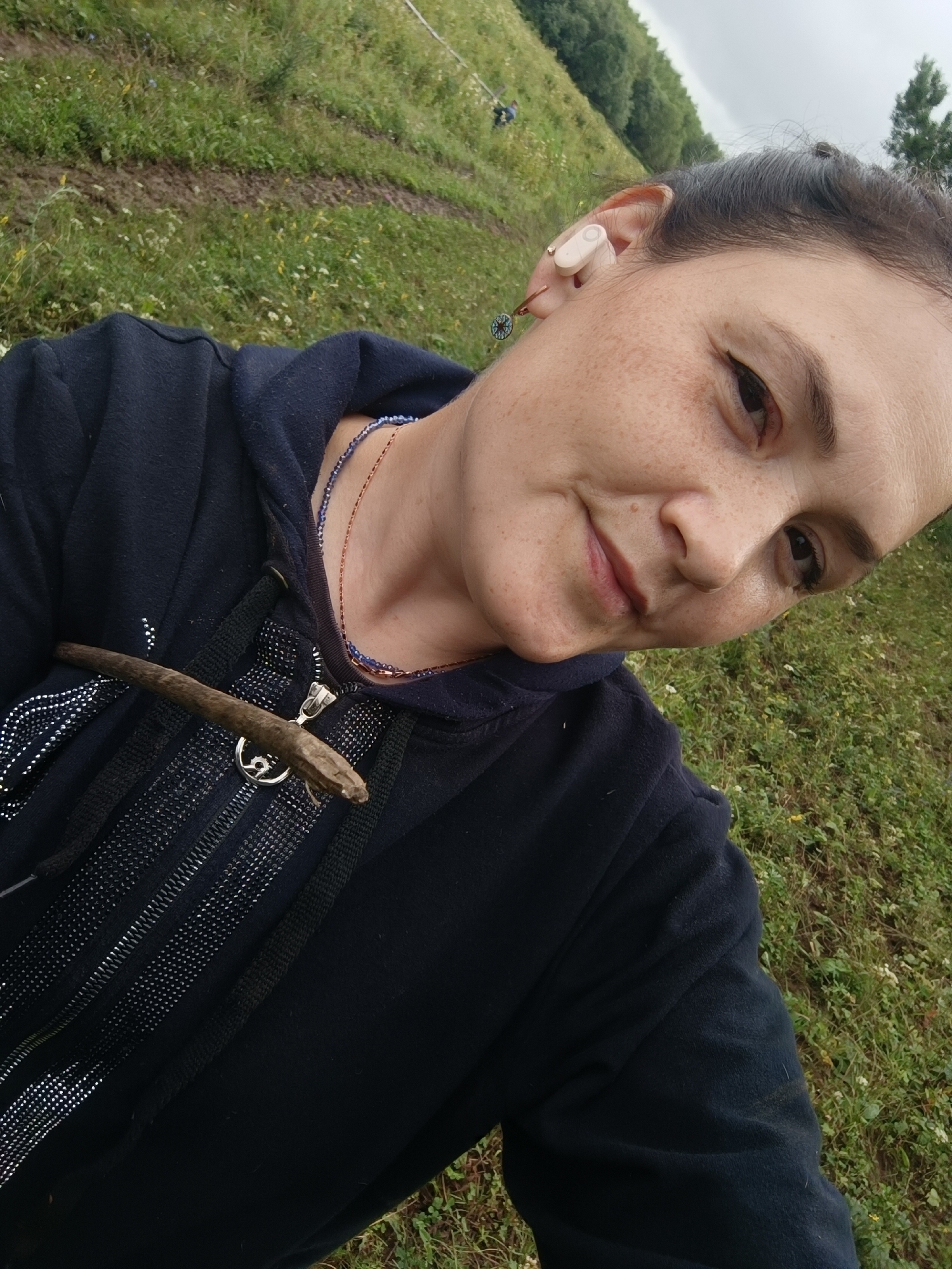 In a low-angle, close-up, selfie-style shot, a woman with a fair complexion, freckles, and dark hair pulled back is captured outdoors. She is wearing a dark blue hooded jacket with a zippered front. A small, brown stick is resting on the left side of her jacket, near the zipper. Two necklaces are visible around her neck, one appears to be made of small blue beads, and the other is a thinner, copper-colored chain. She has a single white earbud in her right ear and a small, circular earring with a dark charm in her left ear. The background consists of a grassy, uneven terrain with some dirt paths and scattered trees in the distance, under an overcast sky. The scene does not provide any information to indicate the location is the city of Karsha, Russia.