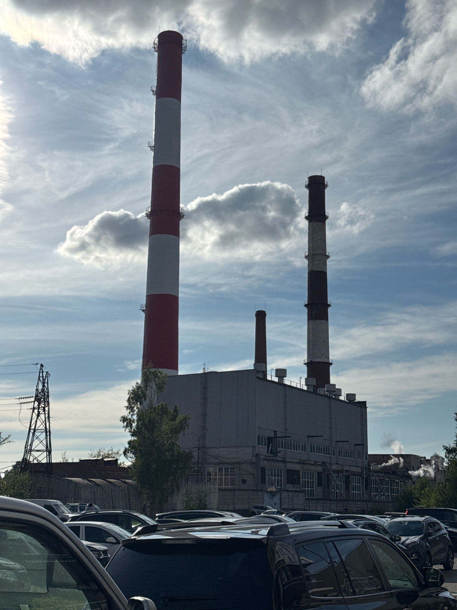 In Ladozhskoe Ozero, Russia, a power plant is visible under a partly cloudy sky. The main structure is a long, gray industrial building with many windows. To the left of the building stands a very tall, red and white striped smokestack, and to the right, a shorter, dark smokestack. In the foreground, numerous cars are parked in rows, suggesting a nearby facility or parking lot. The environment includes industrial infrastructure like a tall transmission tower with wires to the left of the smokestacks and some trees. Wisps of steam or smoke are visible rising from the industrial building in the distance.