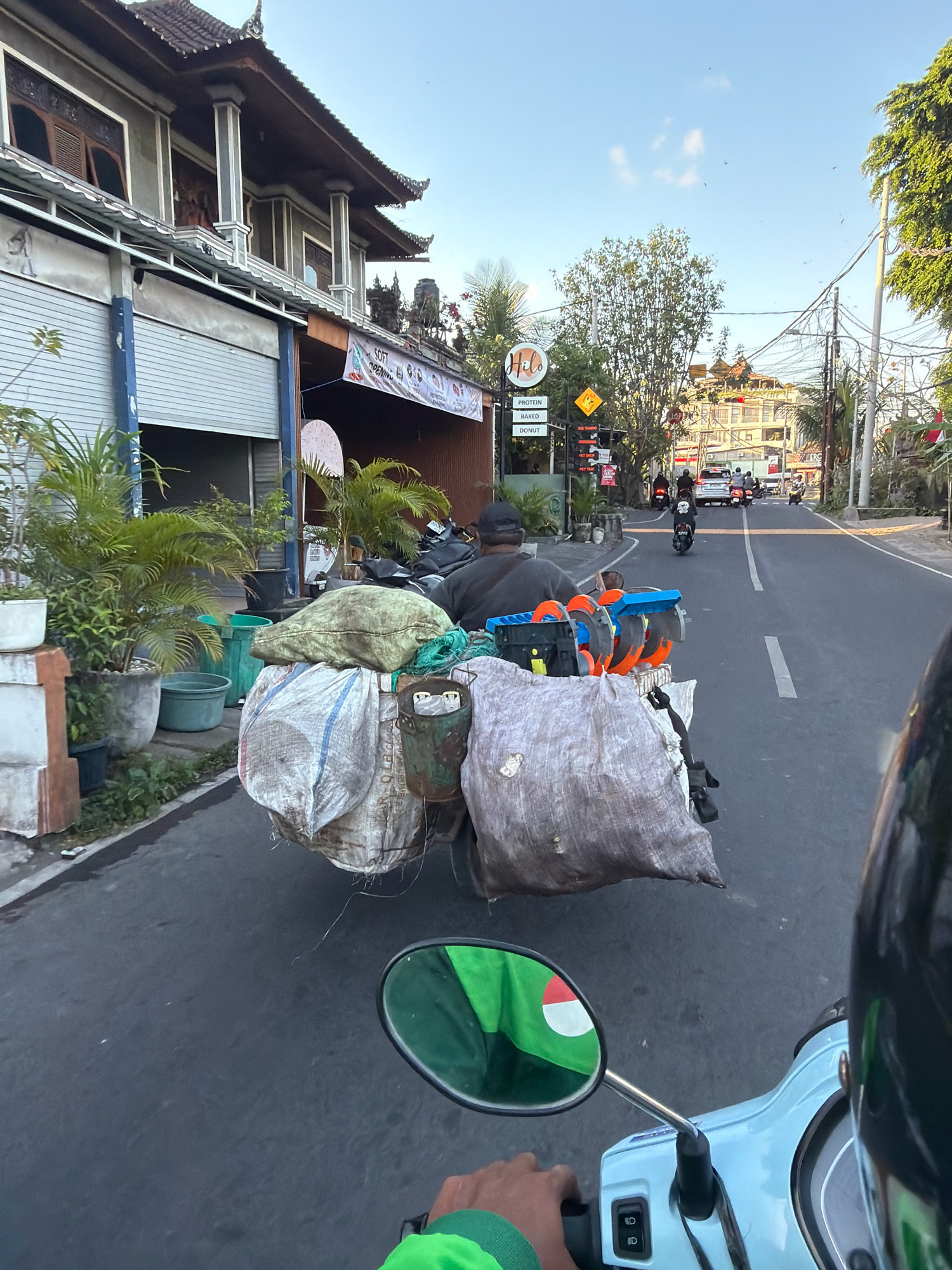 In an outdoor, daytime urban scene, a light blue scooter navigates a paved road with a white line marking the lane. The perspective is from the scooter's rider, looking forward. On the left side of the road, a two-story building with traditional architecture features dark wooden windows and a tiled roof. A metal roll-up door is visible on the ground floor, with potted plants in front. A banner reads "SOFT OPENING" with text below. Further down the road, a sign displays "HILO" in a circle. Multiple other scooters and cars are visible on the road, some moving in the same direction as the viewer, others in the opposite lane.
The scooter in the foreground is carrying a large, overloaded cargo rack. The cargo includes several large, bulging sacks secured to the rack, one appearing to be a rough, woven material filled with light-colored contents, and another made of a lighter fabric with blue and red stripes. Attached to the back of the rack are several orange and blue items, possibly tools or equipment. A person wearing a dark cap and a dark shirt is seated on the scooter, facing forward. A metal bucket is attached to the side of the cargo rack, containing white cylindrical objects.
The environment is sunny, with a clear sky and some scattered clouds. Trees line the right side of the road, and telephone wires are strung overhead. The overall impression is that of a busy street in a Southeast Asian city, possibly Bali, Indonesia, given the architectural style and the presence of scooters as a primary mode of transportation.