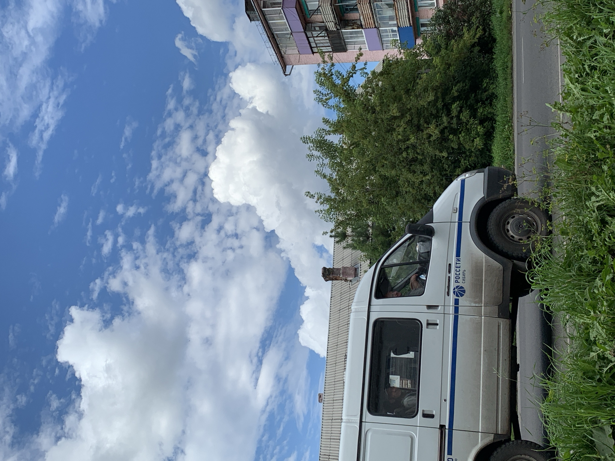A white van with blue stripes is driving on a paved road lined with green grass and weeds. The van has two people inside, one in the driver's seat and another visible in the passenger seat. The van has text on its side that says "РОССЕТИ СИБИРЬ" and a logo. Behind the van, there is a gray-roofed building with a brick chimney, and further in the background, a multi-story pink building with balconies. Trees are present between the buildings. The sky is blue with scattered white clouds. The image appears to be taken from a side angle, looking towards the front of the van. The scene is consistent with a residential or commercial area in Prokopyevsk, Russia.