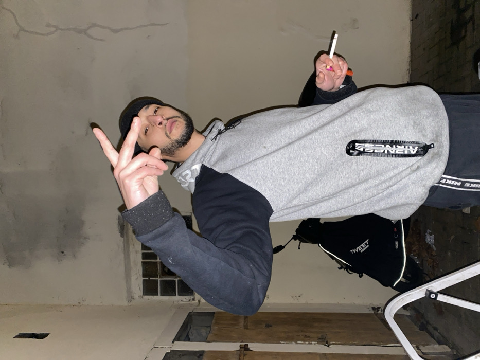 A man in a black beanie, gray and black hoodie, and black pants poses indoors, holding a cigarette and making a peace sign. He has a beard and mustache. The background shows damaged walls and a window. A pair of black shoes is visible on the right, alongside what looks like a ladder. The lighting is dim and the picture appears rotated.