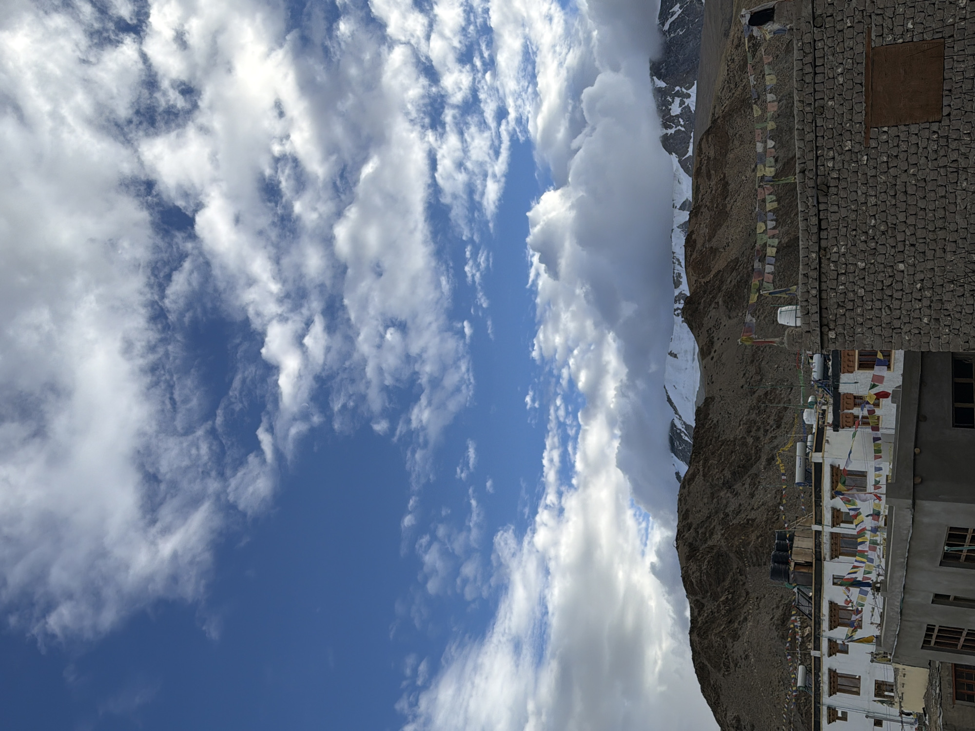 The image shows a cloudy sky over the city of Karzok, India. The sky is a deep blue with white clouds scattered across it. In the foreground, there are buildings with prayer flags strung between them. The buildings are made of white stucco with wooden window frames. Behind the buildings, there are mountains. The mountains are brown and rocky, with snow-capped peaks. The overall scene is one of peace and serenity.