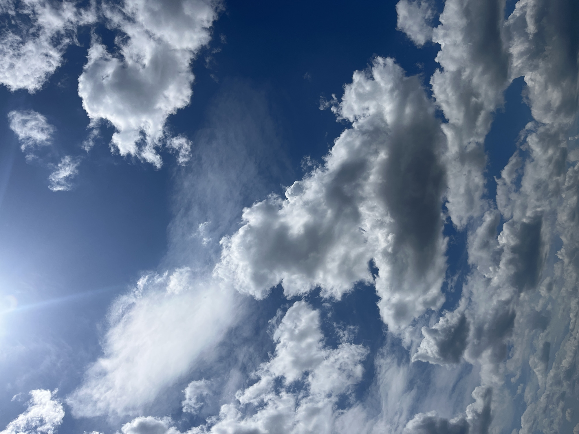 The image depicts a daytime sky with cumulus clouds against a blue backdrop. The sun is visible in the lower-left corner, creating a bright glare. The clouds vary in size and density, some appearing fluffy and white, while others are darker with gray undertones. No people, subjects, or actions are directly observable in the scene, nor any objects besides the clouds themselves. The environment is limited to the sky. It is not possible to infer a location in the city of Varna, Bulgaria, from the image alone.