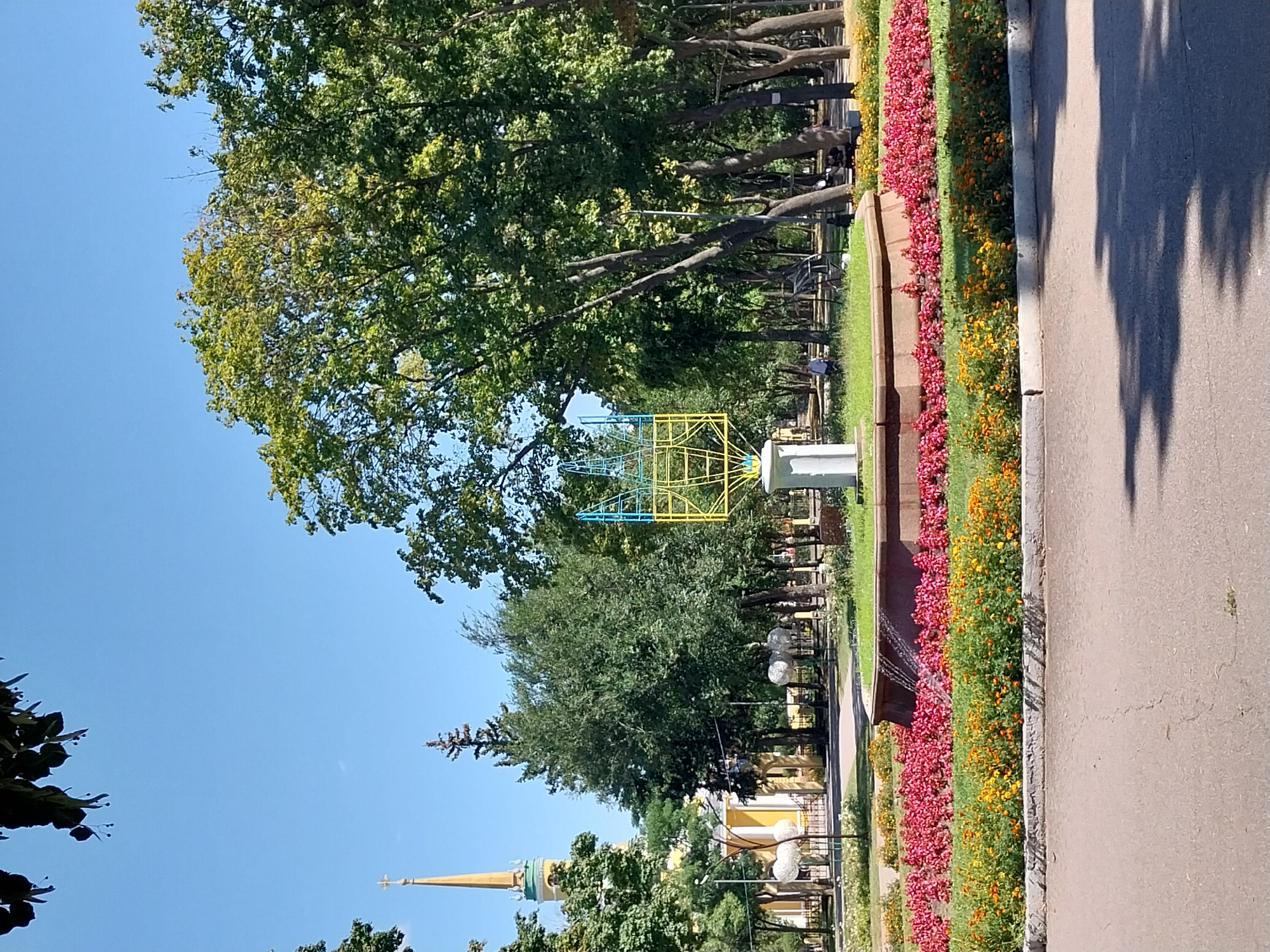 The image depicts a sunny day in a park in Dnipro, Ukraine. In the foreground, a paved path with shadows from overhead trees runs horizontally across the bottom of the frame. Beyond the path is a flowerbed with two distinct layers of flowers: a lower row of yellow and orange flowers and an upper row of vibrant pink flowers. Behind the flowerbed, a tiered fountain is visible, with water cascading down one side into the fountain's basin, which is surrounded by grass.
In the middle ground, a sculptural installation, a geometric structure made of yellow and blue metal frames, stands on a white pedestal. Behind the sculpture, a large fountain basin with red flowers and green grass is prominent. Further back, several large, mature trees with lush green foliage line a park pathway. To the left, a yellow building with a tall, slender spire topped with a cross is partially visible, hinting at a church or religious institution. Several smaller trees and decorative garden elements are scattered throughout the park. A few people are visible in the background, appearing as small figures walking or sitting in the park, indicating a public space in use. The sky is clear and blue, characteristic of a warm summer day. The overall impression is a well-maintained public park with a mix of natural and artistic elements.