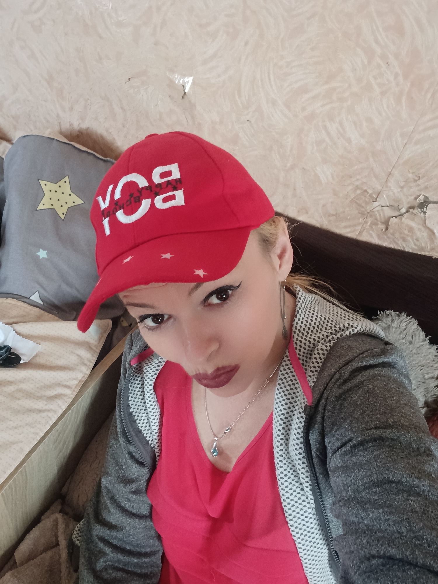 The media file shows a close-up, high-angle selfie of a young woman wearing a red baseball cap with white lettering that appears to say "BOY" with some additional obscured text. She has blonde hair, dark eyeliner, and is wearing a pink top and a grey hooded jacket. A silver necklace with a light blue pendant is visible. She is seated, with a grey pillow featuring a yellow star visible behind her left shoulder. The background appears to be a textured wall and a dark surface, possibly a couch or bed. The lighting suggests it is daytime. The woman's expression is a playful pout. The image was taken in Chernihiv, Ukraine.