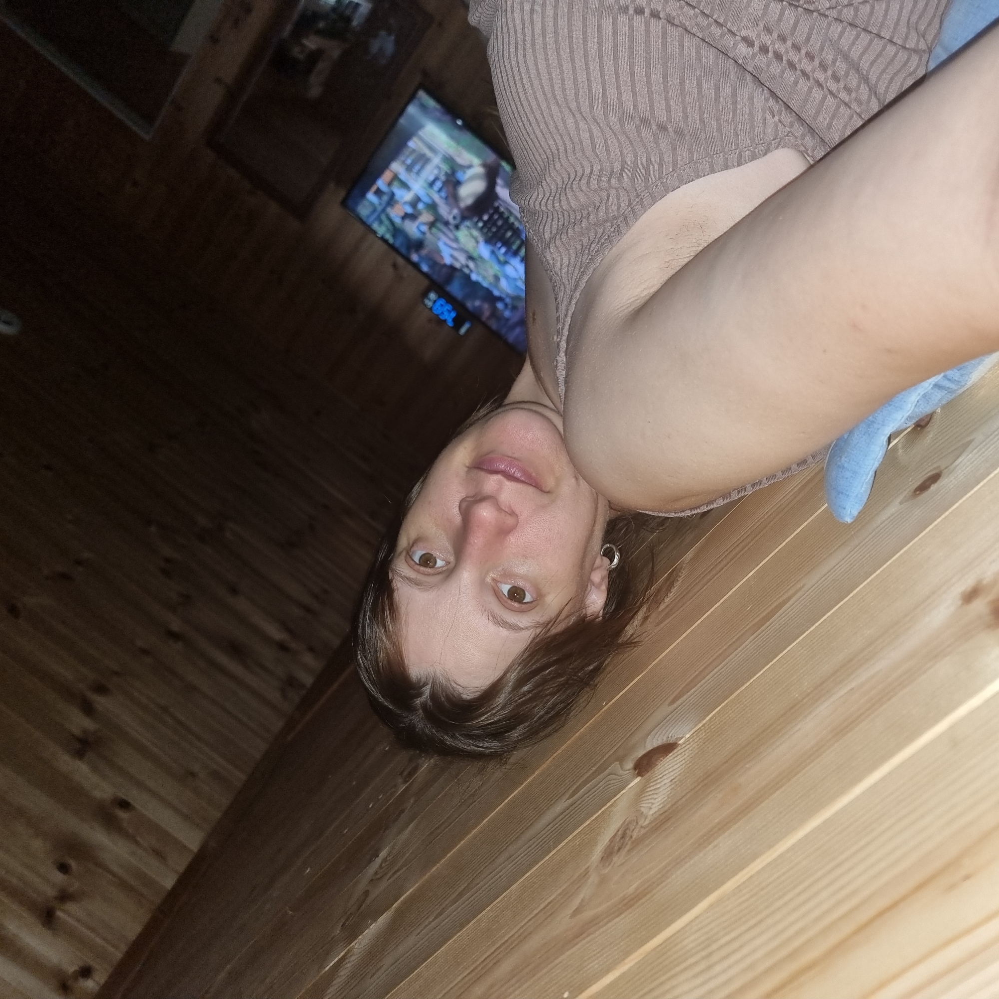 The image shows a close-up, tilted shot of a woman lying down on a wooden surface, likely a wall or floor paneling made of light-colored wood planks. The woman has brown hair and is looking towards the camera with a slight smile. She is wearing a light brown, ribbed garment. In the background, a television screen is visible, displaying an image that appears to be from a video game or animated content. A digital clock showing "7:59" is also visible on the television stand. The overall setting appears to be indoors, with the wood paneling suggesting a rustic or cabin-like atmosphere. There are no explicit indications of the location being in Dunnae-myeon, South Korea.