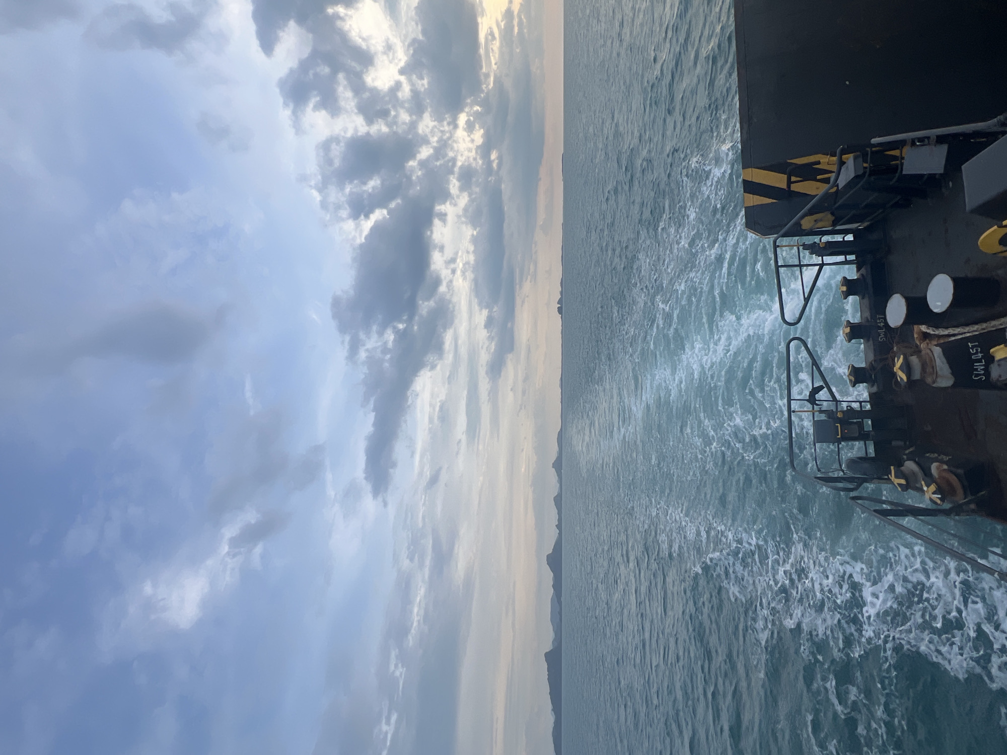 An over-the-shoulder view from the rear of a dark vessel looking out at a calm ocean and a hazy sky. The wake of the boat stretches out in a white foamy trail towards a distant horizon where a series of dark, low-lying islands are silhouetted against the pale sky. The sky itself is filled with soft, greyish-blue clouds that are breaking up to reveal patches of lighter sky. The deck of the vessel is visible in the foreground, featuring mooring bollards and a railing. The overall scene suggests a tranquil journey at sea, possibly at dawn or dusk.