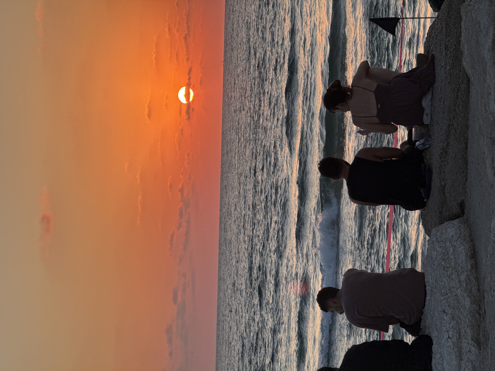 A group of people are sitting on rocks on the shore of the Mediterranean Sea in Tel Aviv, Israel, watching the sunset. The sky is a vibrant orange and pink, with a few wispy clouds. The sun is a bright orange orb, partially obscured by a cloud. The sea is a deep blue, with waves gently lapping at the shore. The people are silhouetted against the sunset, and their faces are not visible. One person on the left is wearing a black shirt, another is wearing a grey t-shirt, and a woman on the right is wearing a purple dress. There are some plastic containers and a black flag on a pole in the foreground. The overall mood of the image is peaceful and serene.
