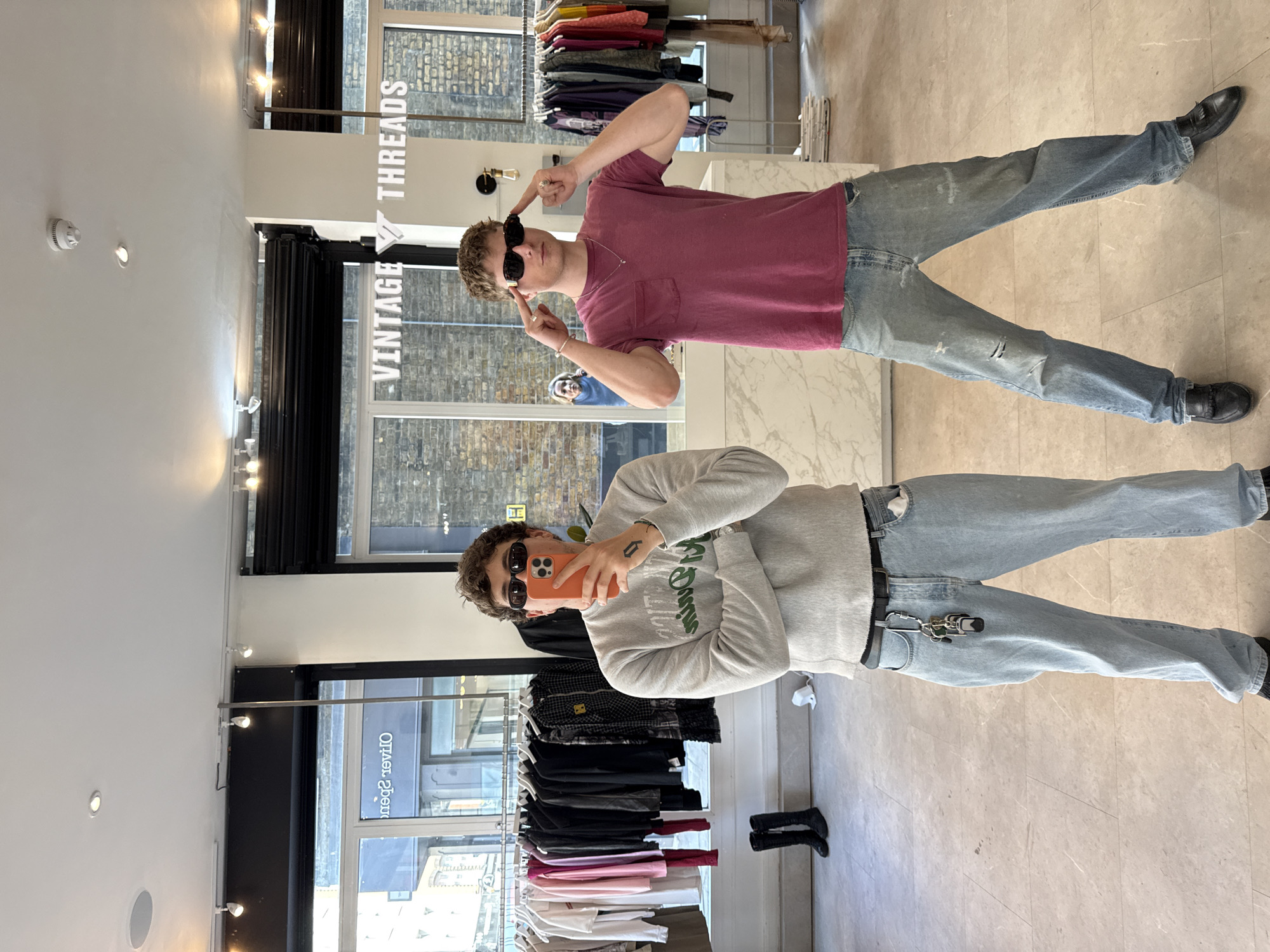 The image displays two young men posing casually inside a clothing store, likely "Vintage Threads," in London, United Kingdom. The store features a modern interior with light-colored walls and a tiled floor, illuminated by overhead track lighting and natural light from large windows.
On the left, one man takes a mirror selfie with an orange phone. He wears dark sunglasses, a grey crew-neck sweatshirt with green text (partially obscured), light-wash jeans, a black belt with a prominent keychain attached, and black shoes. His dark hair is styled casually.
On the right, another man stands posing playfully, also wearing dark sunglasses and pointing at them with both index fingers. He has short, light brown hair and is dressed in a maroon short-sleeved t-shirt and light-wash, distressed jeans, paired with black loafers. A small silver necklace is visible around his neck.
In the background, behind the man in maroon, a prominent white sign with "VINTAGE THREADS" written in bold, capital letters is mounted on a column, reinforcing the store's identity. Various racks of clothing, including t-shirts and shirts in different colors, are visible throughout the store. Through the large windows, reflections of brick buildings and a reversed sign that reads "Olivier Spencer" can be seen, hinting at an urban setting. The overall atmosphere is relaxed and candid, capturing a moment of fun within the retail environment during what appears to be daytime.