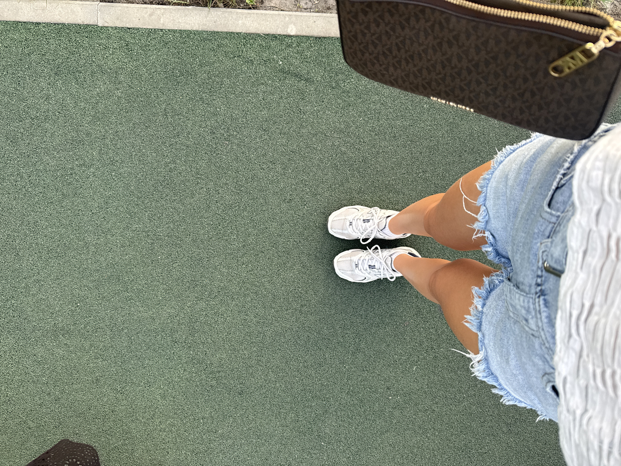 Here is a forensic description of the image:
The image is a high-angle shot of a person standing on a green, textured surface, possibly a rubberized or synthetic material. The person is wearing white sneakers, frayed denim shorts, and a partially visible white top. A brown Michael Kors bag is partially visible in the upper right corner of the frame. The background is primarily the green surface, with a hint of a concrete edge visible in the upper left corner. The specific location is unknown. No actions or interactions are discernible.