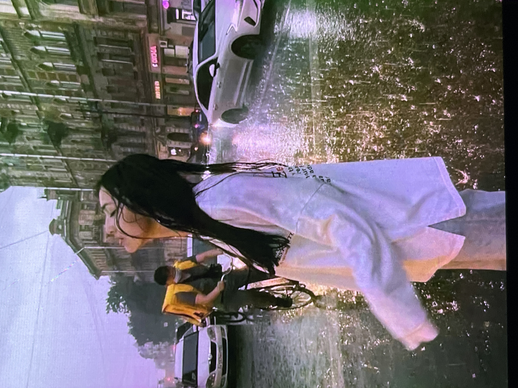 The image depicts a rainy street scene in the city of Cadek, Indonesia. In the foreground, a person wearing a white long-sleeved shirt and light-colored pants stands facing away from the camera, with their head tilted back and eyes closed. Their wet dark hair is spread out behind them. To the left, a delivery person wearing a yellow insulated bag on their back and a face mask rides a bicycle through the rain. In the background, an older white car drives through the wet street, creating splashes of water. Several multi-story buildings with ornate architecture line the street. The sky is overcast and a hazy purple hue.