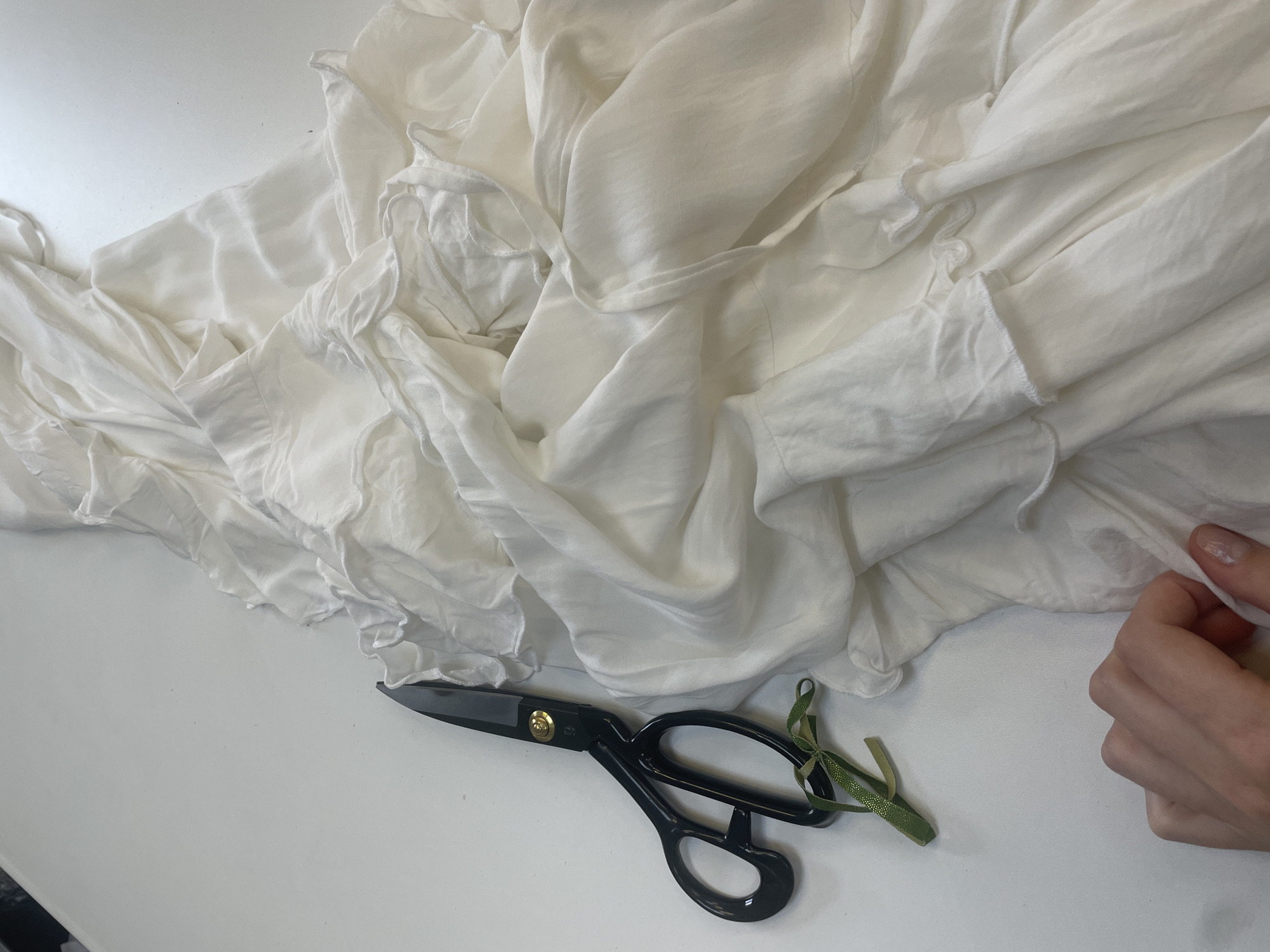 A pair of black-handled scissors with a gold screw and a green ribbon bow are on a white surface next to a rumpled pile of off-white fabric. The fabric has ruffles and appears to be a light material such as silk or chiffon. A person's hand with manicured nails is reaching into the fabric on the right side of the image. The lighting is bright and casts soft shadows. The scene appears to be a workspace, possibly for sewing or crafting. There is no indication of a specific city or location.