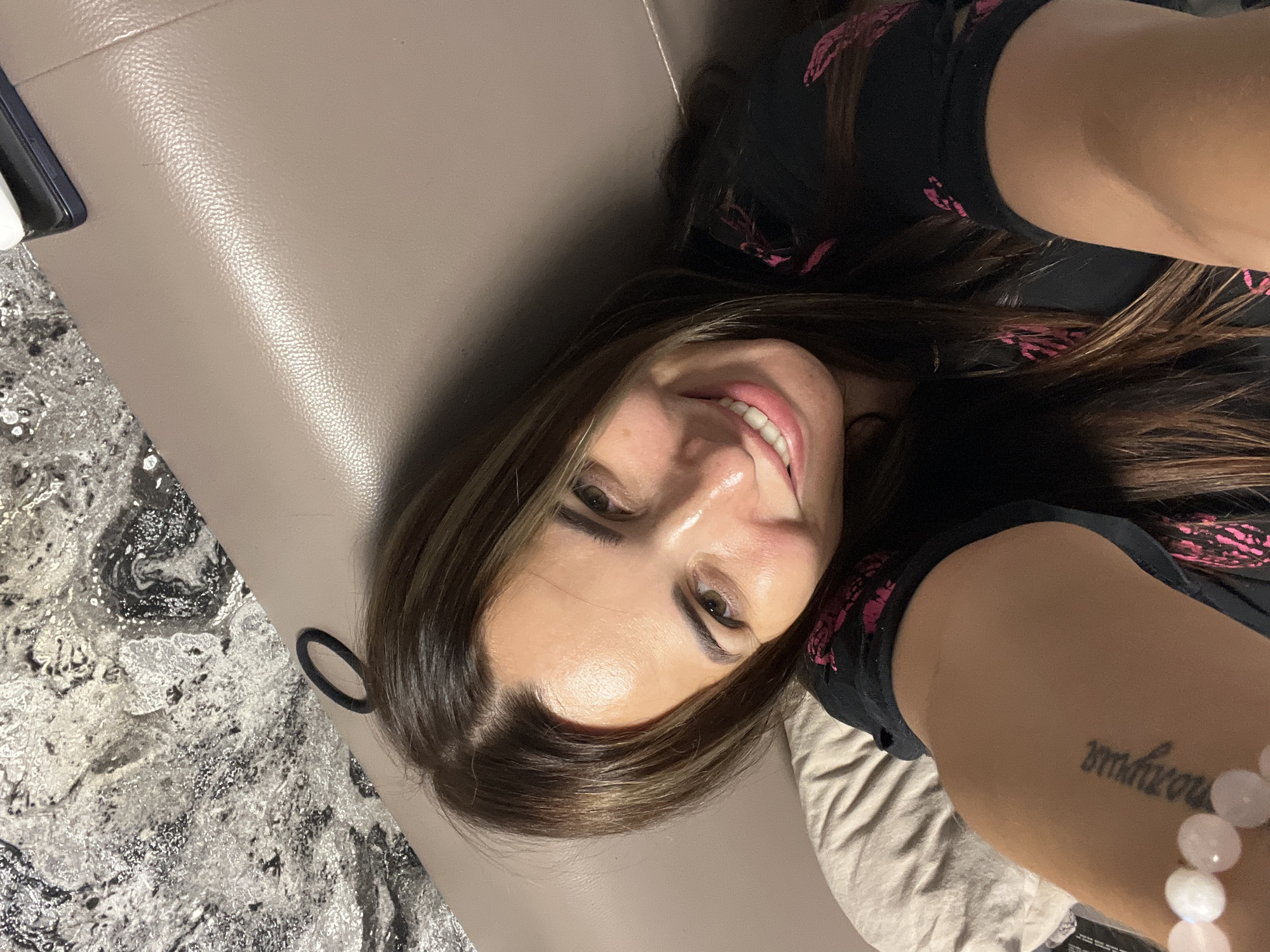 The image shows a woman lying down, taking a selfie. She has long, dark brown hair with highlights and is wearing a black t-shirt with a pink design. She is smiling with her teeth showing. She has a tattoo on her arm that appears to be in Greek script. There is a light-colored surface behind her, possibly a sofa or bed, and a patterned floor or rug in the foreground. The lighting suggests it could be indoors. The woman appears happy and relaxed.