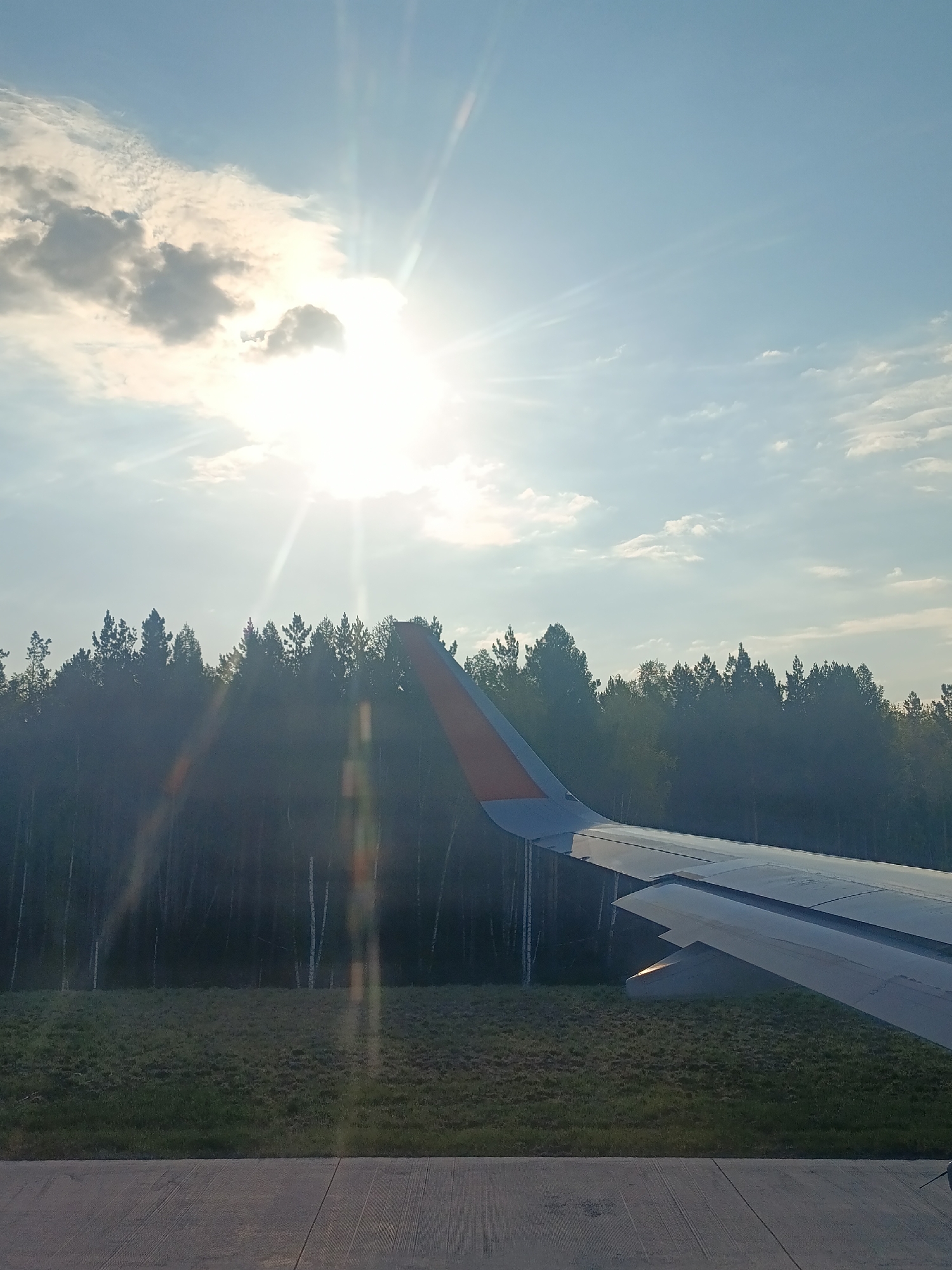 View from an airplane window. The sun is high in the sky, partially obscured by clouds. Below is a view of a forest and a grassy field. The wing of the airplane is visible in the foreground. The ground appears to be an airport runway. The location is Yelovoye, Russia, based on the presence of birch trees in the forest.