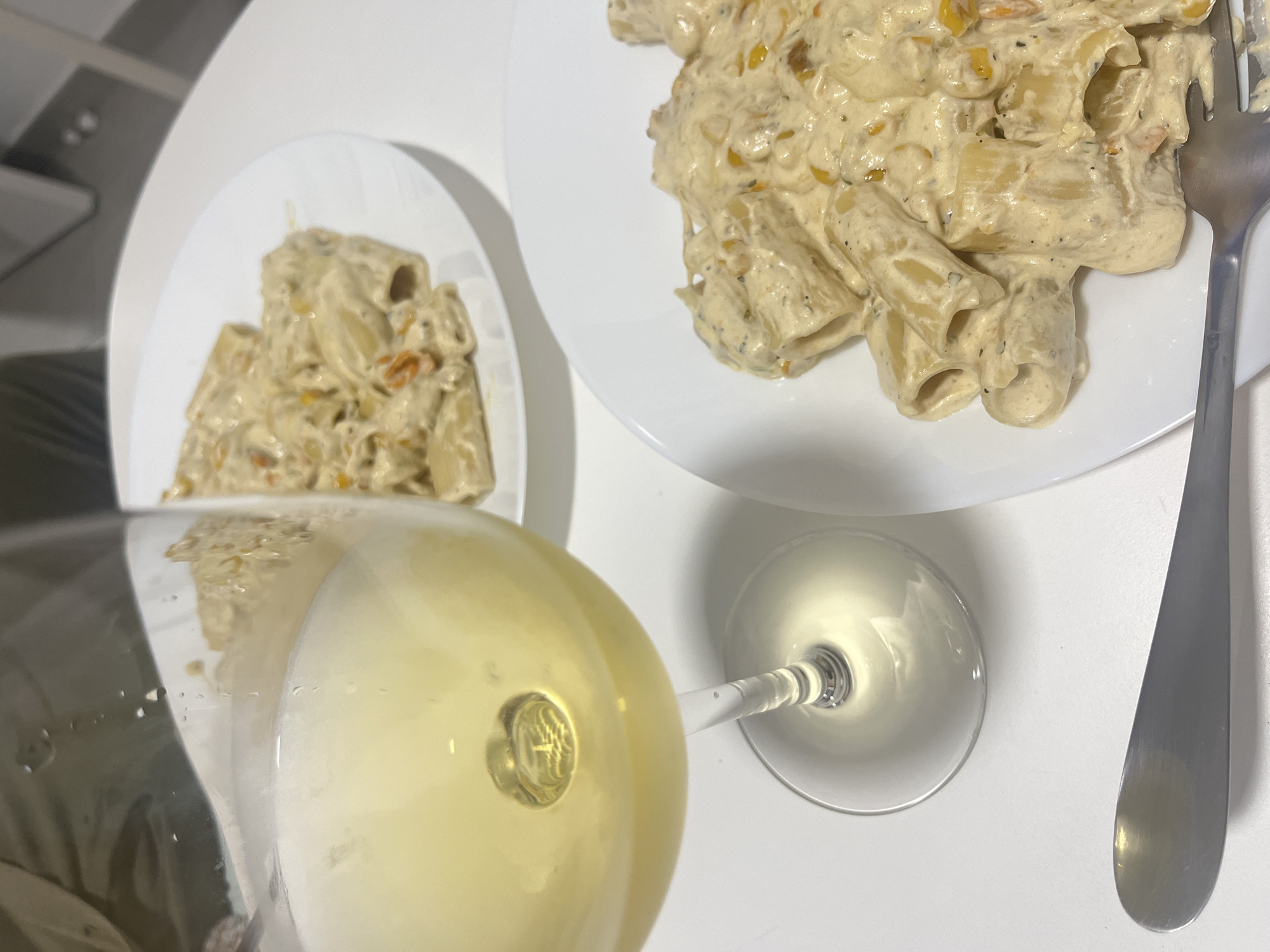 The image presents a still-life composition, presumably indoors, featuring a dining setting. Two white plates hold portions of pasta, appearing to be rigatoni, covered in a creamy sauce. Adjacent to the plates is a silver fork. A wine glass filled with a light-colored liquid, likely white wine, is also visible. The background is plain and white. There is nothing to indicate this scene is located in Mogán, Spain. No people, interactions, or specific environmental details are discernible.